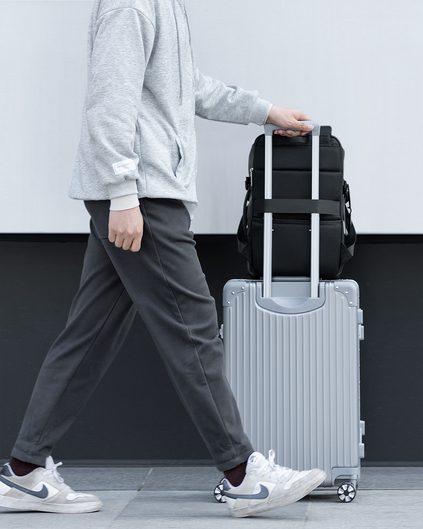 Person mit grauem Kapuzenpullover zieht Koffer und Rucksack vor weißem Hintergrund.