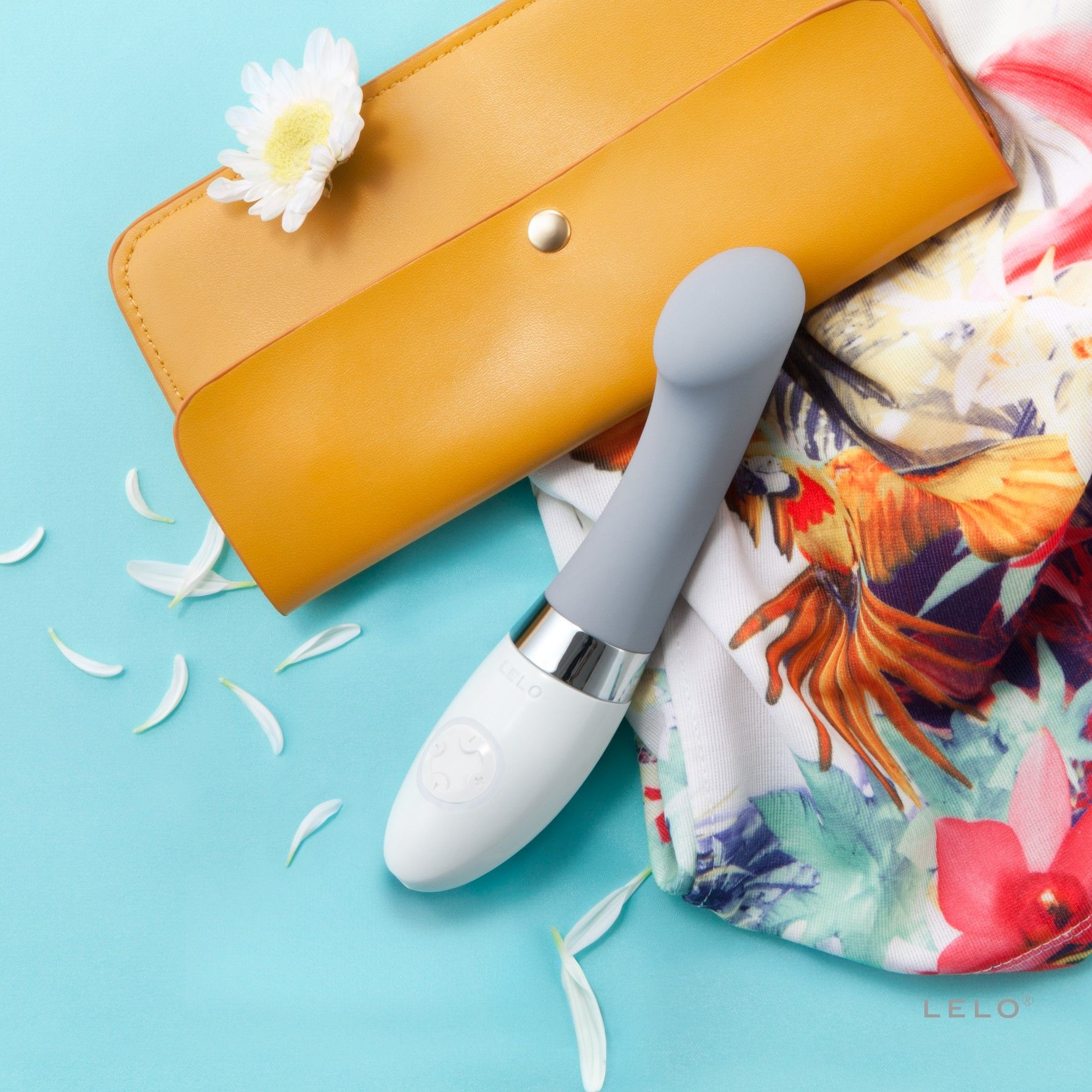 Biała i szara zabawka erotyczna na niebieskiej powierzchni. Obok, żółty portfel i tkanina we wzory kwiatowe.