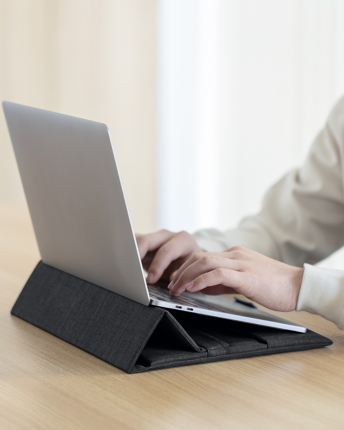 Eine Person tippt auf einem Laptop, der auf einem dunkelgrauen Laptopständer auf einem Holztisch steht.