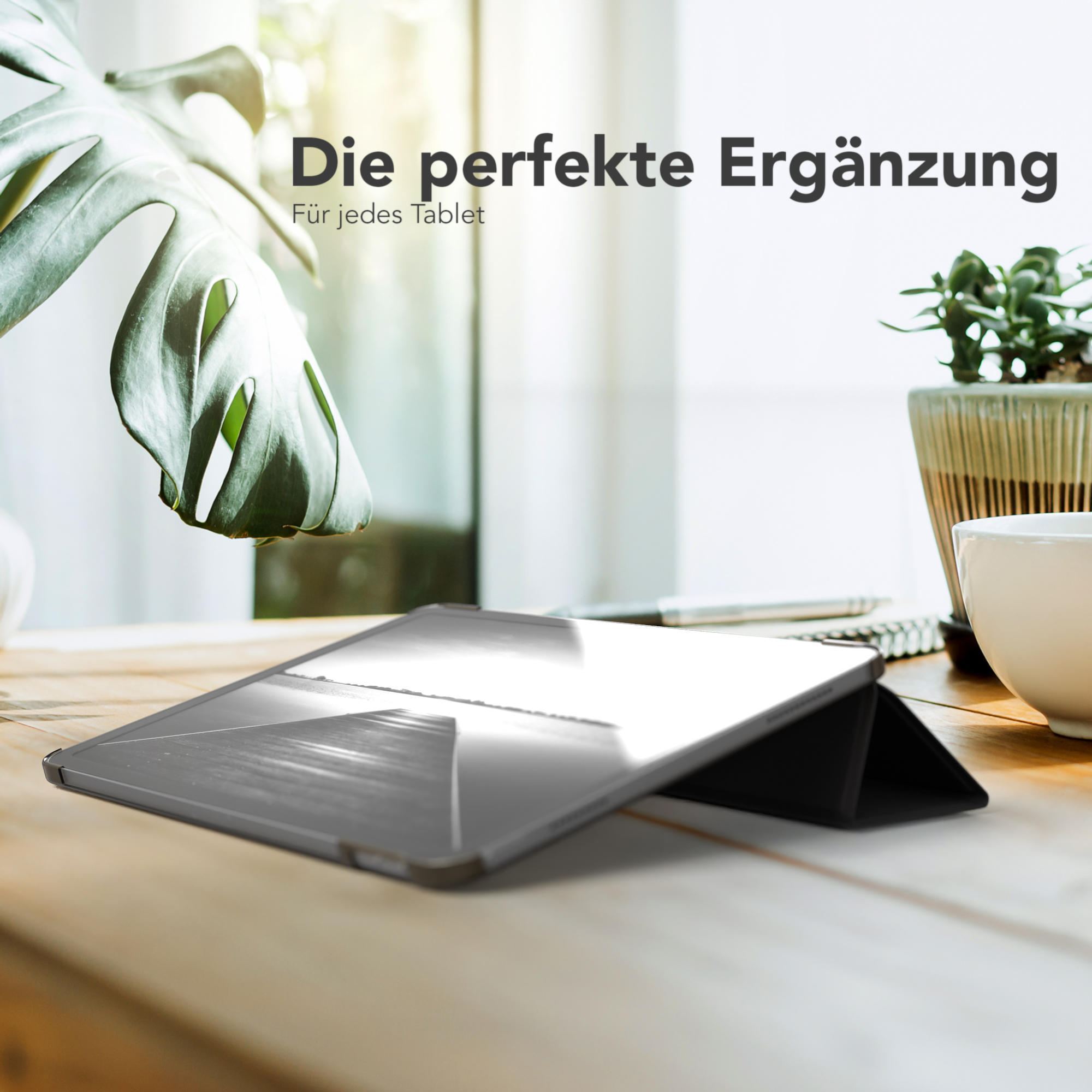 Tablet mit Hülle auf einem Schreibtisch. Grüne Pflanze und eine weiße Tasse im Hintergrund. Helles Fensterlicht.