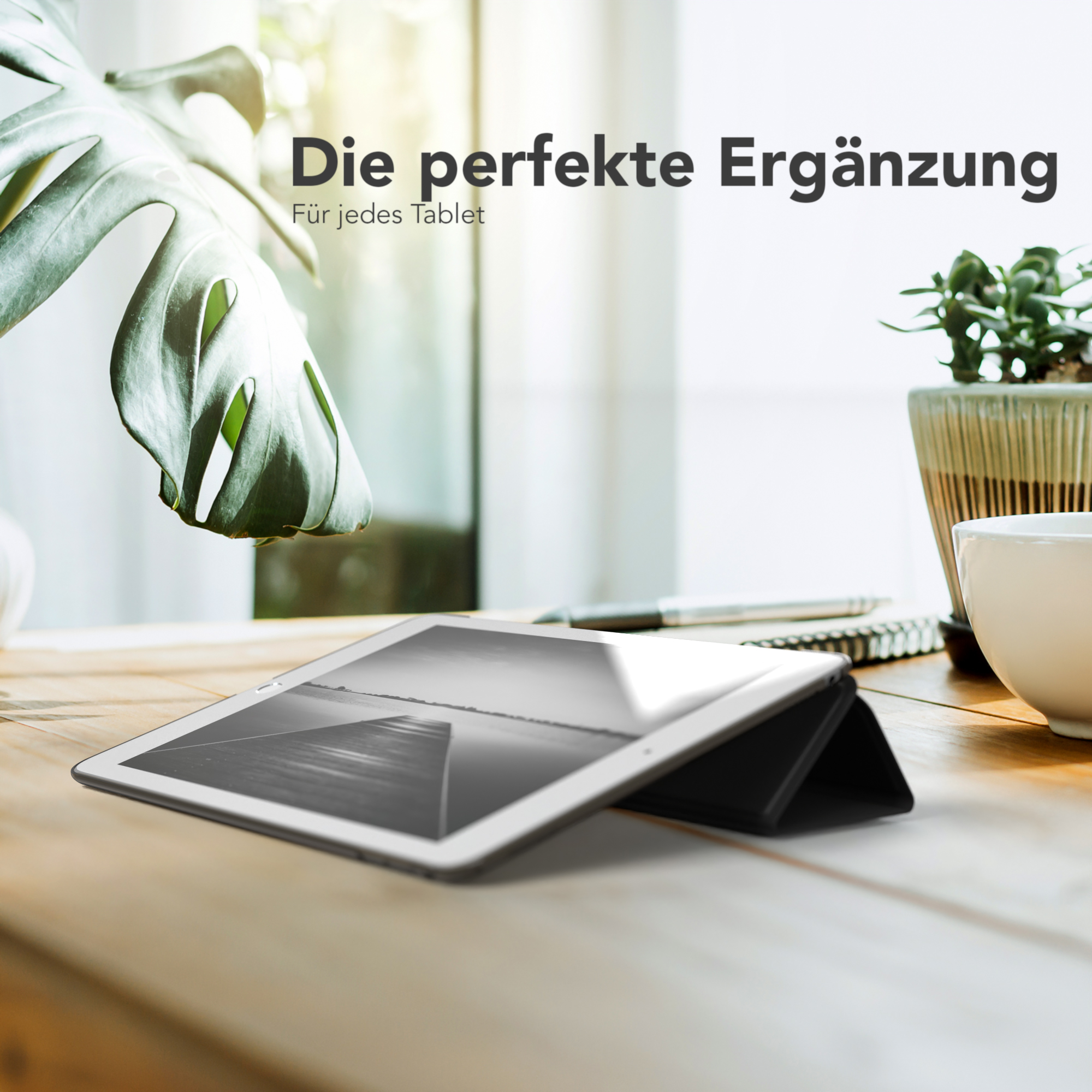 Ein Tablet in einer schwarzen Hülle auf einem Holztisch. Eine Pflanze und eine Tasse sind im Hintergrund.
