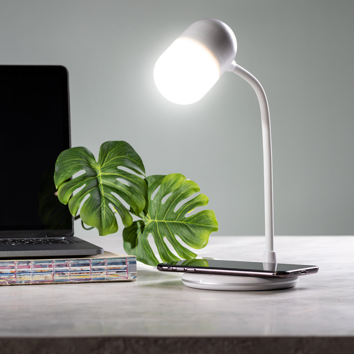 Una lámpara de escritorio blanca con luz brillante, una planta, un portátil y un teléfono sobre una superficie blanca.