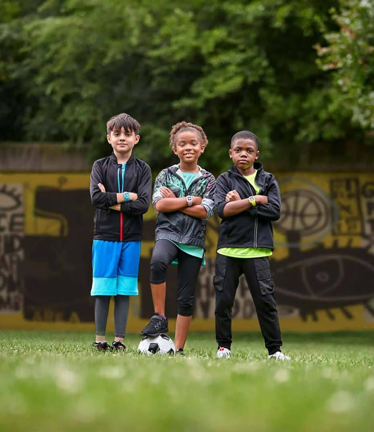 Troje dzieci w dresach stoi na boisku. Jedno dziecko trzyma piłkę nożną.