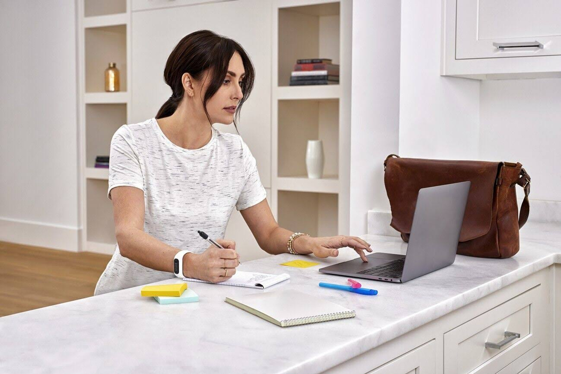 Donna che lavora su un laptop, scrive su un quaderno, con una borsa in pelle vicina.