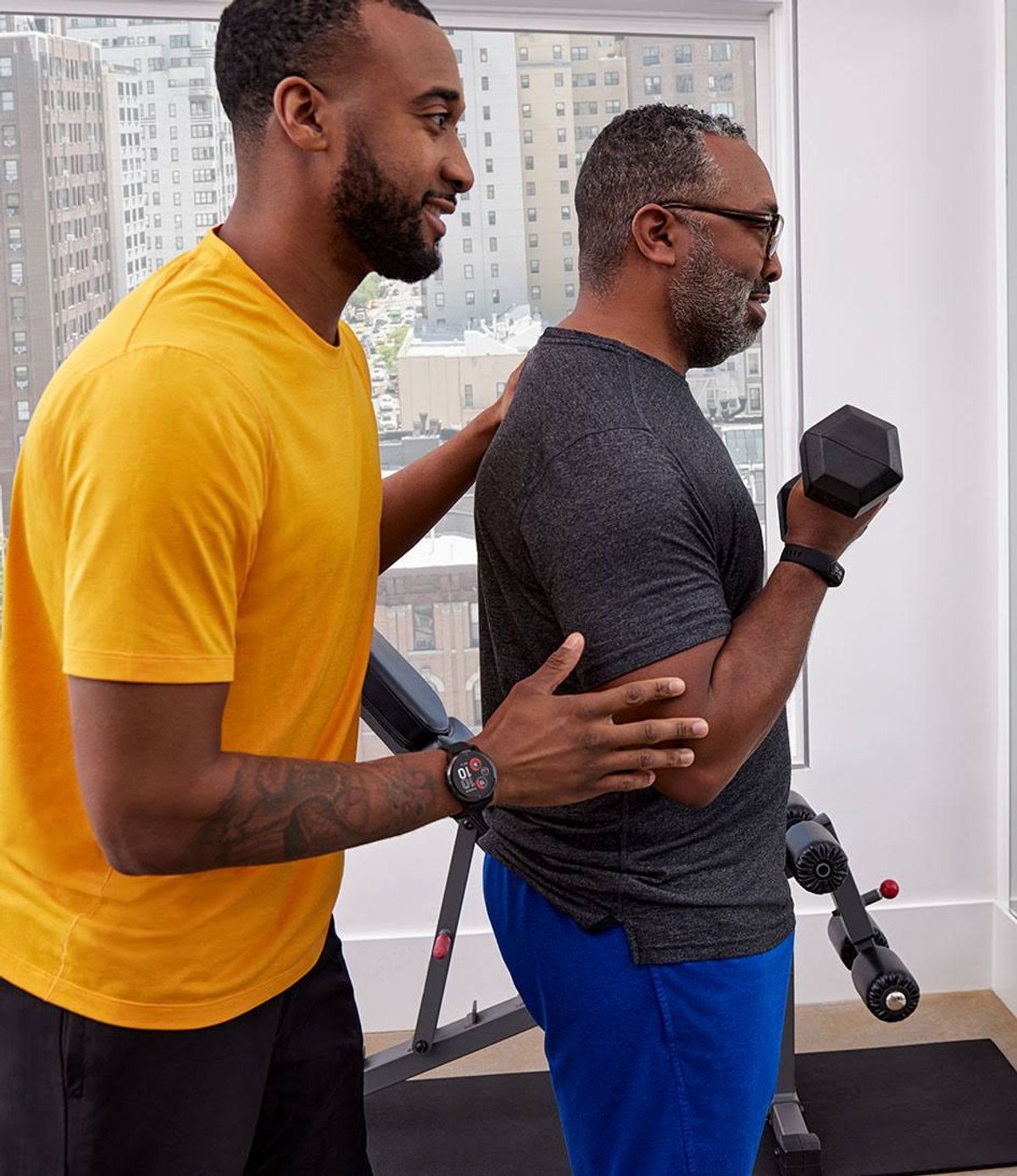 Due uomini in palestra. Uno assiste l'altro con un esercizio con i manubri, braccio esteso.