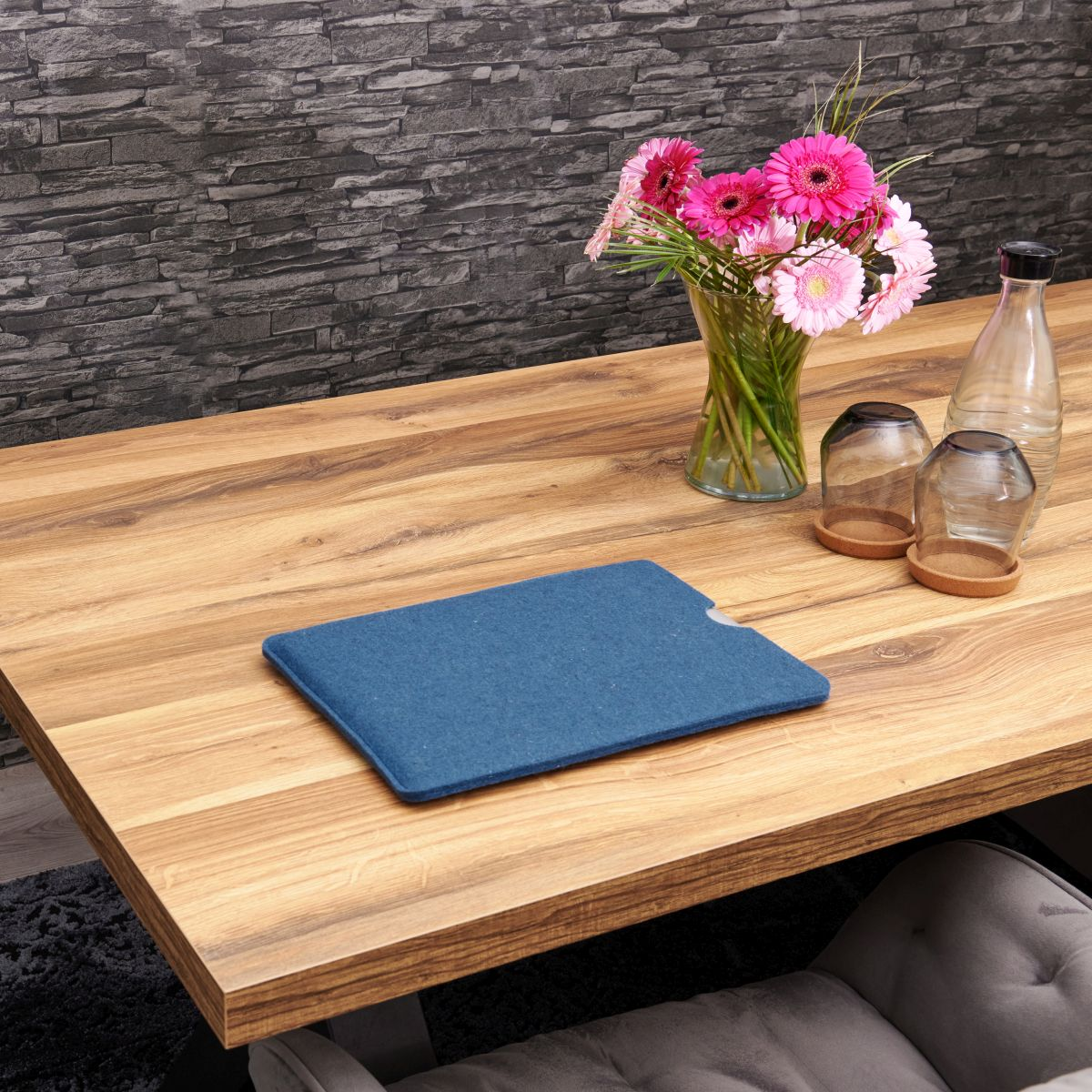 Holztisch mit Blumen, Gläsern und einer blauen Hülle. Dunkler Steinwandhintergrund.