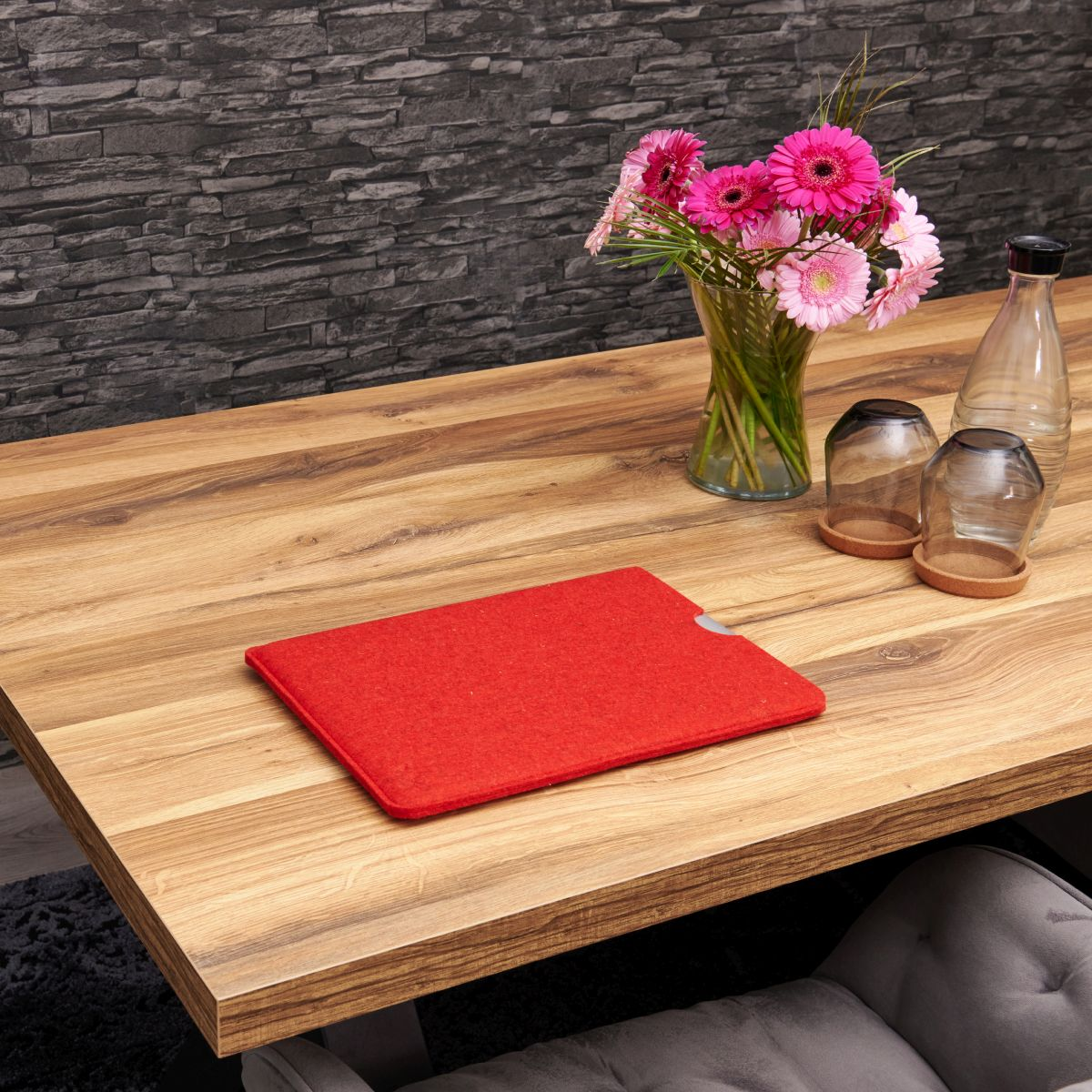 Rote Filz-Laptop-Hülle auf einem Holztisch mit Blumen und Gläsern und grauem Hintergrund.