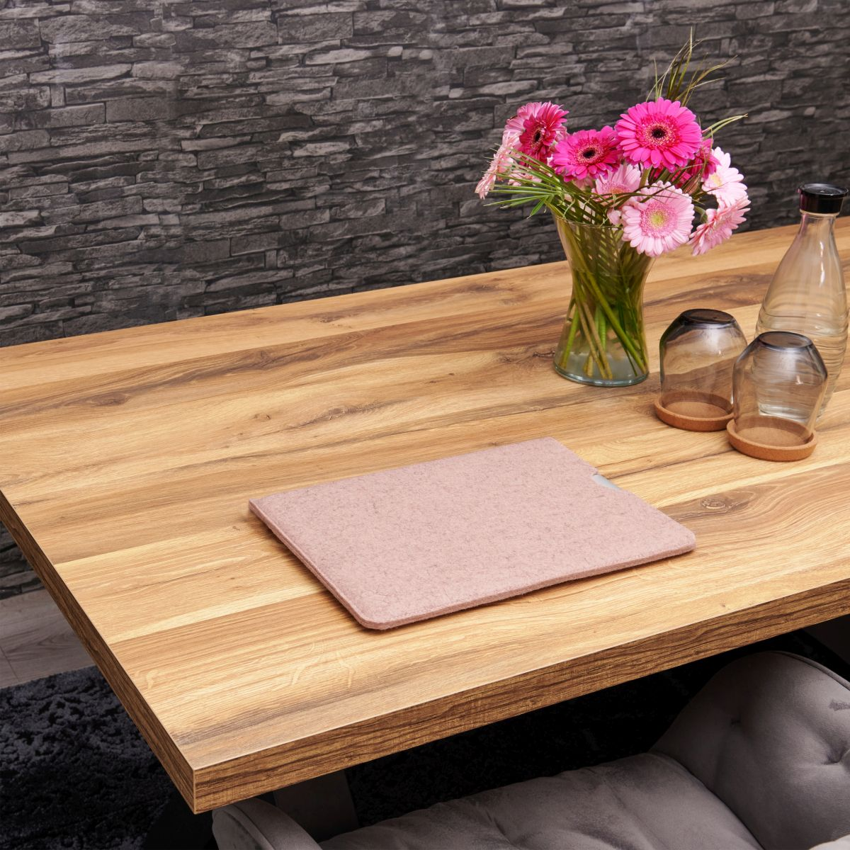 Ein Holztisch mit einem rosa Filzpad, Blumen in einer Vase und Gläsern. Eine dunkelgraue Backsteinmauer ist der Hintergrund.