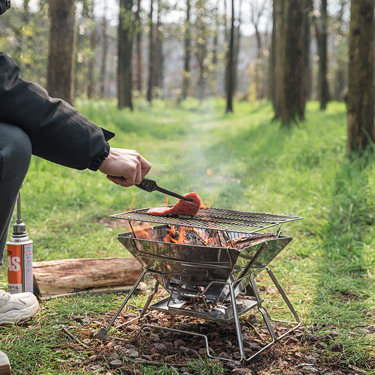 Ein Grill auf einem niedrigen Metallständer mit Feuer und Fleisch. Die Hand einer Person mit Zange arbeitet.