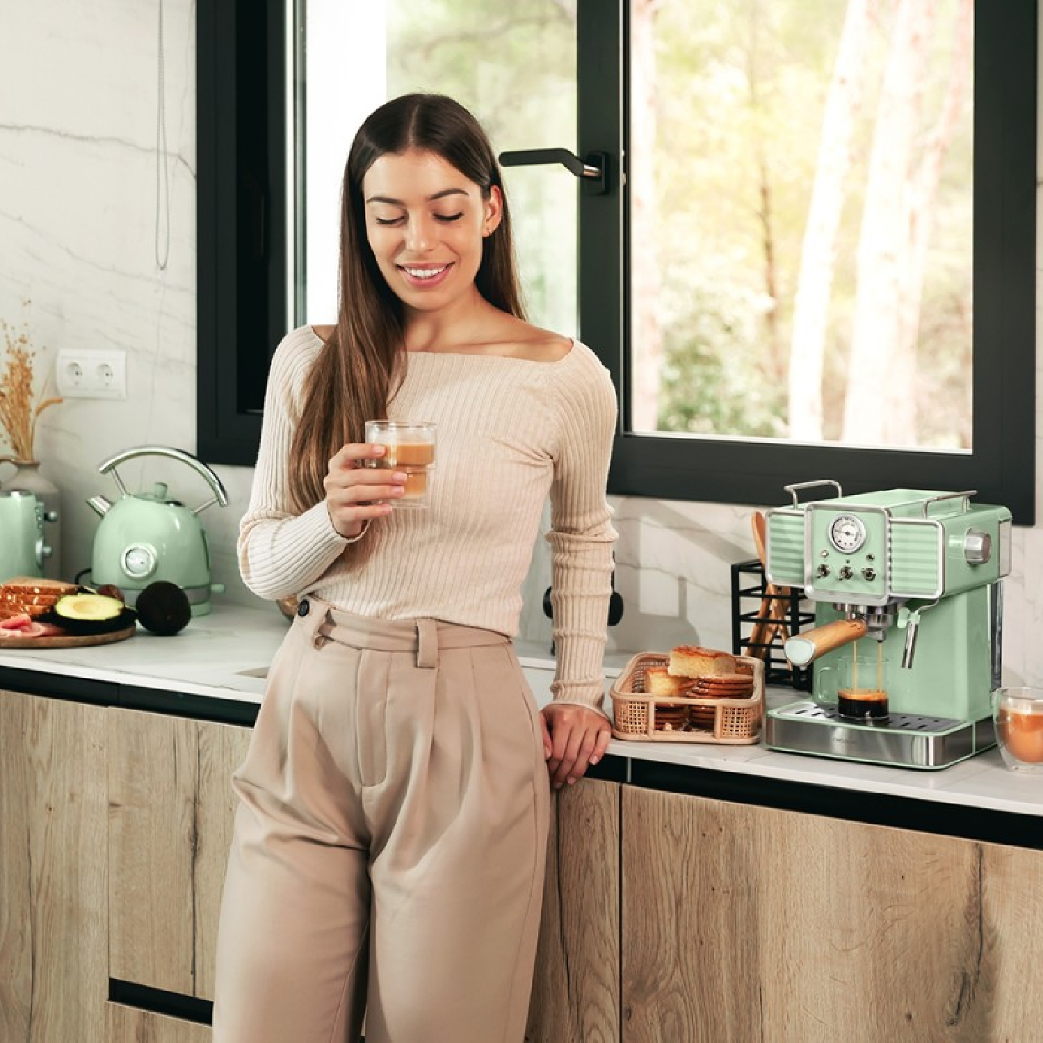 Una donna in cucina con una macchina per il caffè, che tiene in mano un bicchiere di bevanda.