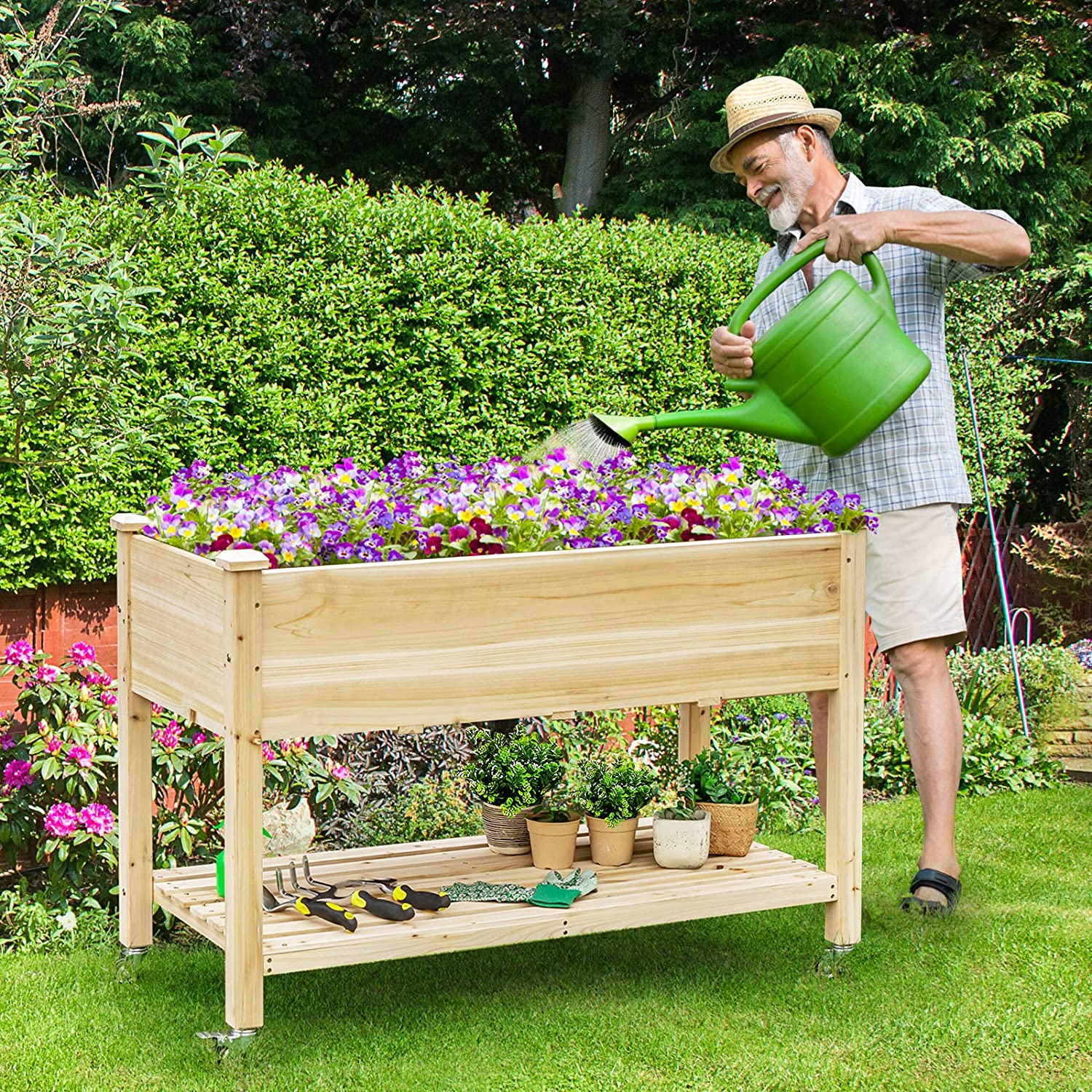 Ein älterer Mann gießt Blumen in einem hölzernen Pflanzkasten auf einem rollbaren Ständer im Garten.