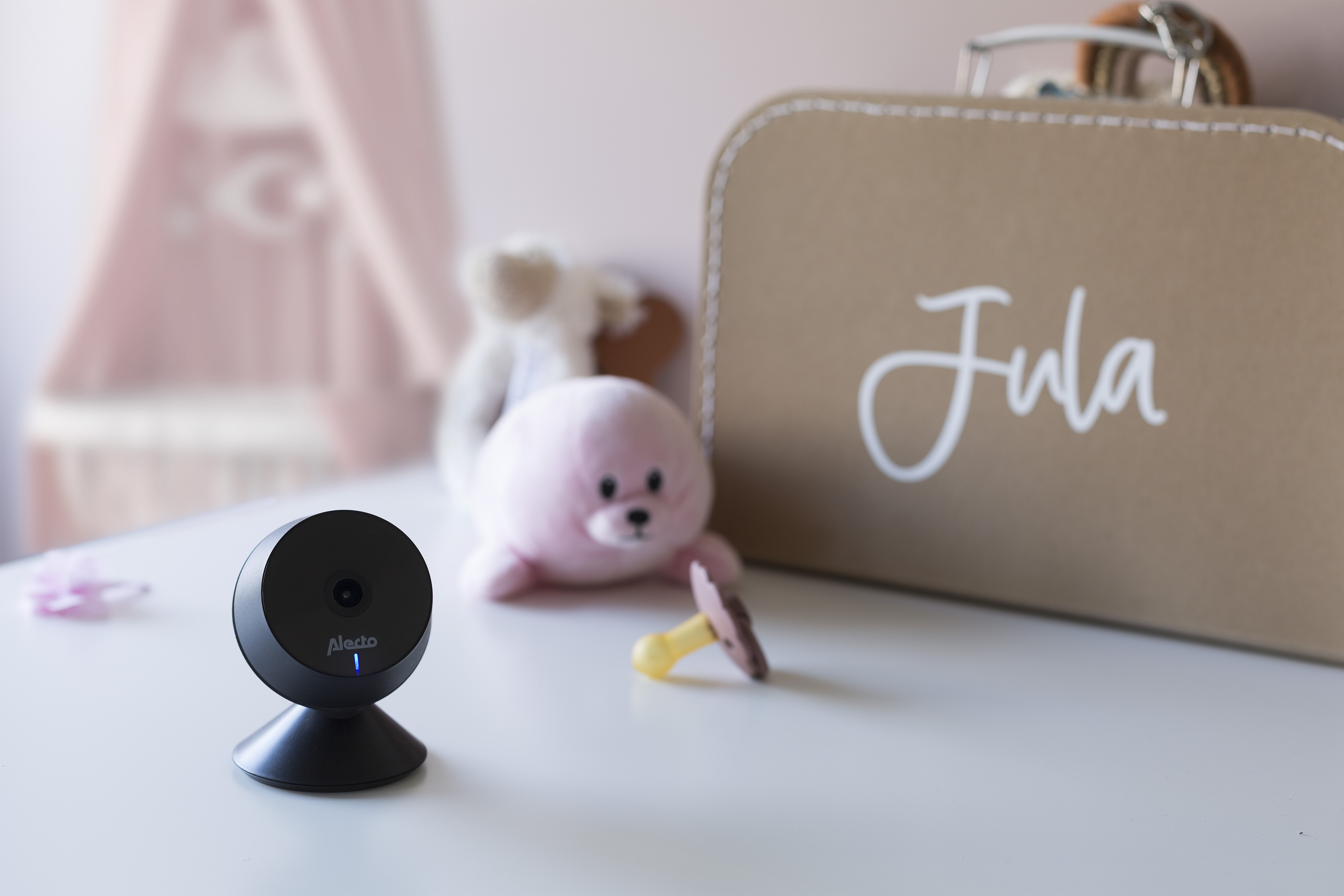 Schwarzer Babyphone auf weißem Tisch. Rosa Bär und brauner Koffer mit dem Namen 'Jula'.