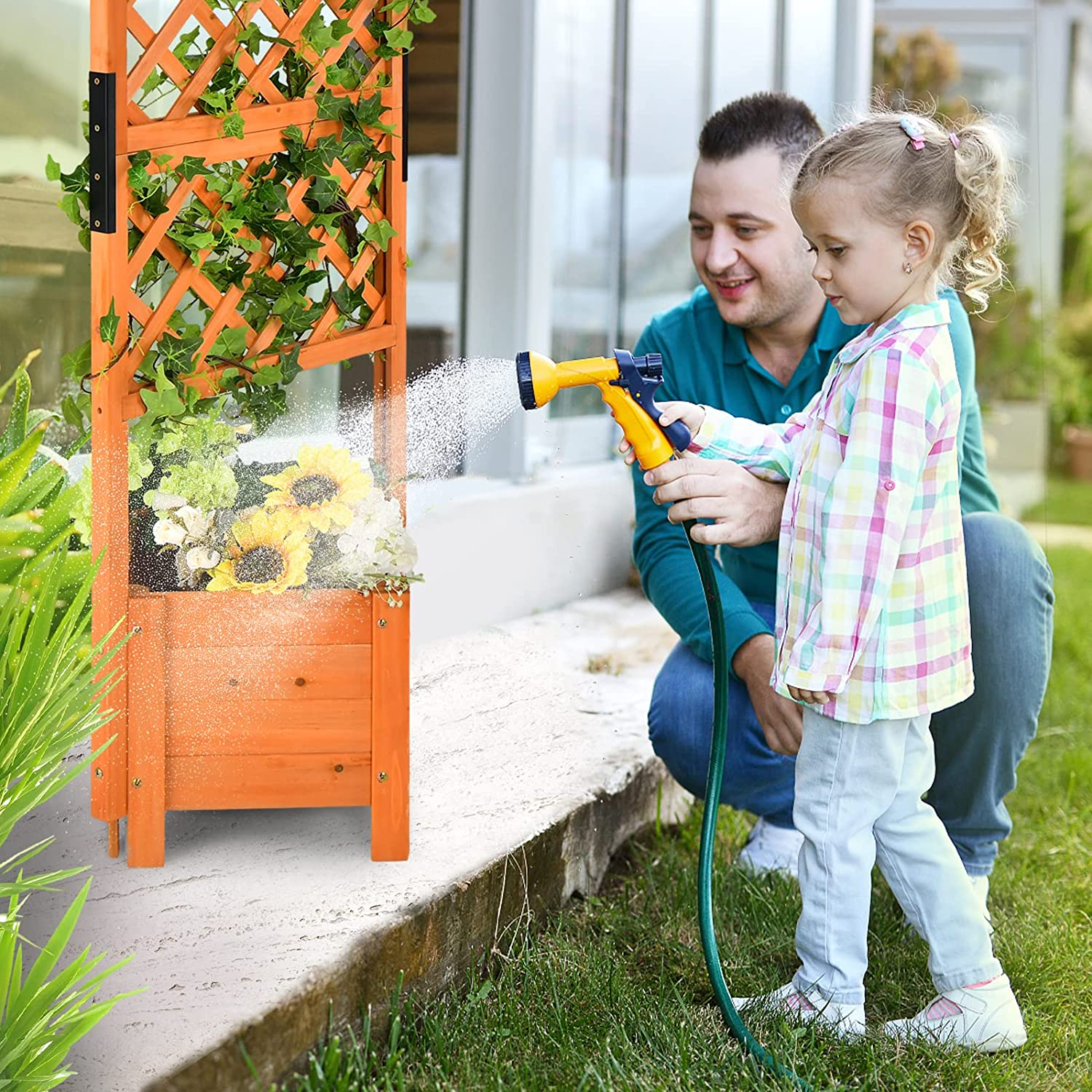 Ein Kind und ein Mann gießen Blumen in einer hölzernen Pflanzkasten mit einem grünen Spalier.