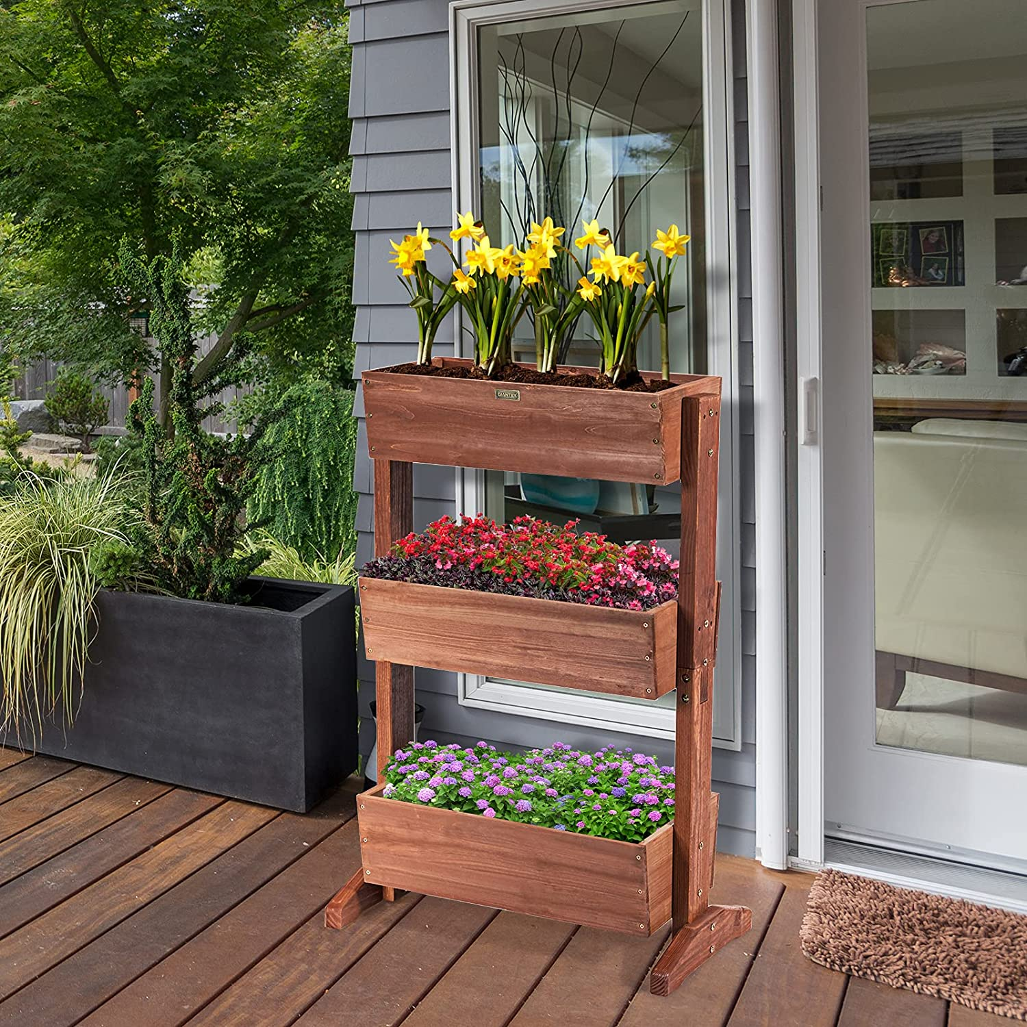 Ein dreistufiger Holzpflanzer mit gelben Narzissen, roten und lila Blumen. Es steht auf einer Holzterrasse.