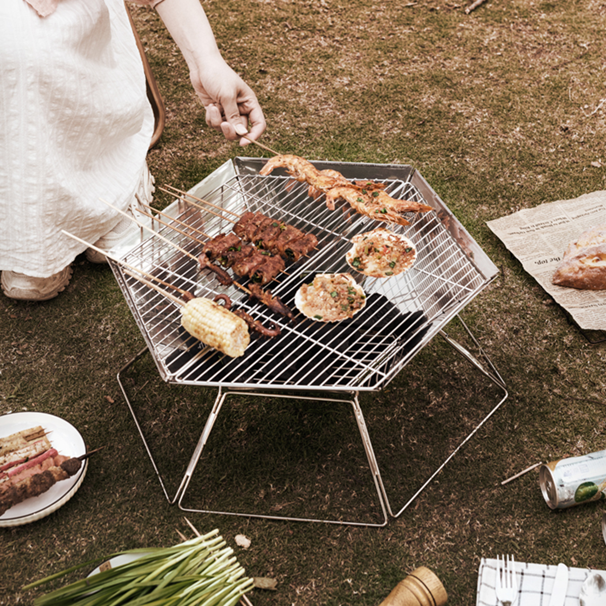 Eine Person grillt Essen auf einem sechseckigen Grill. Essen umfasst Spieße, Mais und Meeresfrüchte auf Gras.