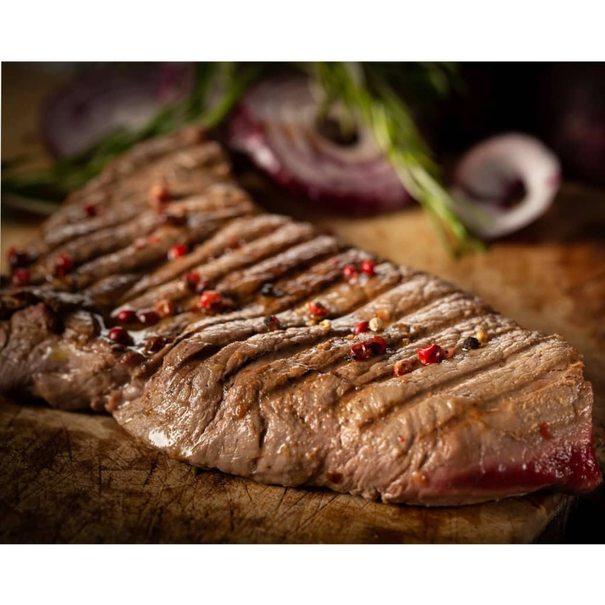 Bistecca alla griglia condita con grani di pepe rosa, su un tagliere di legno con cipolla rossa.