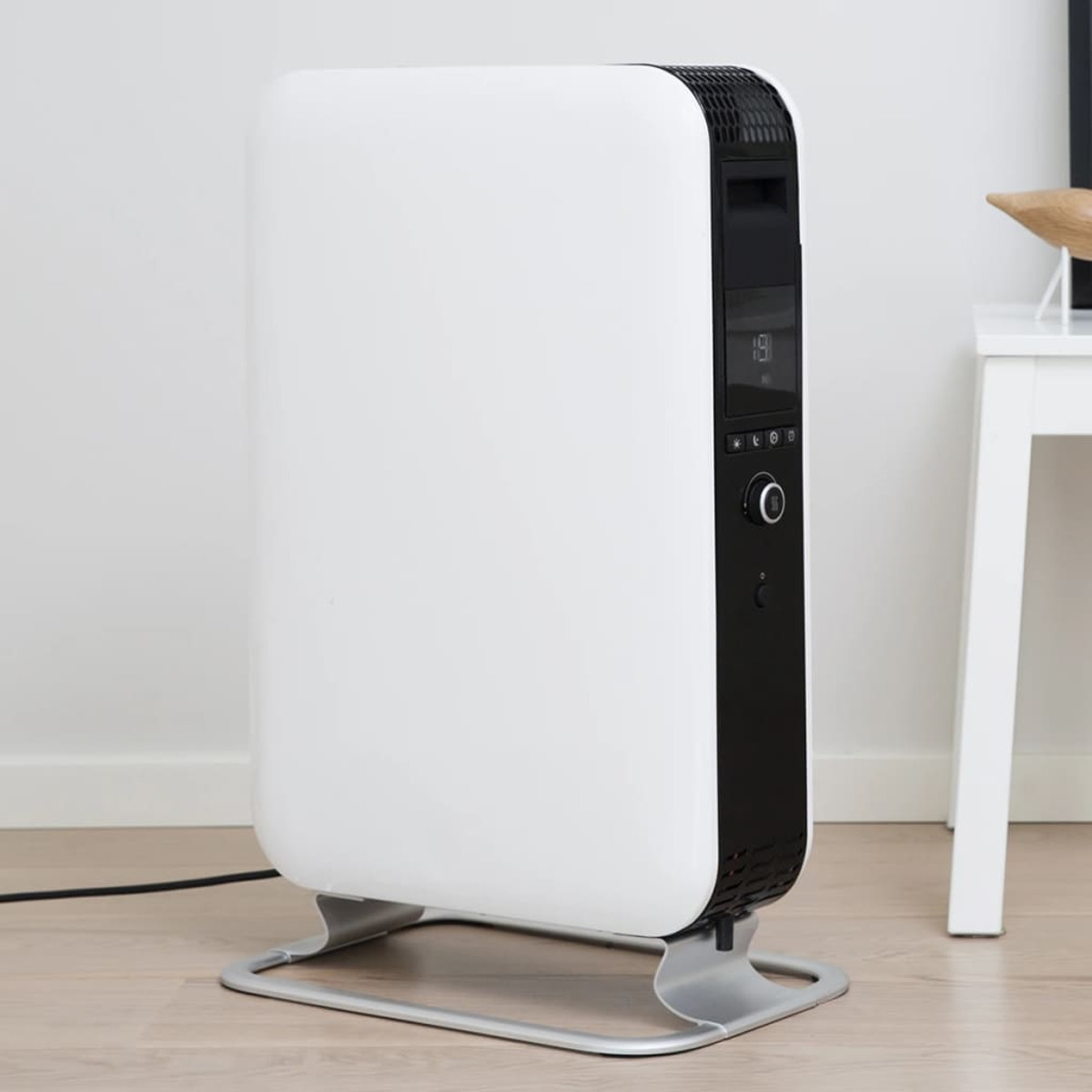 Biało-czarny grzejnik elektryczny na srebrnej podstawie, z wyświetlaczem cyfrowym. Tło białej ściany.