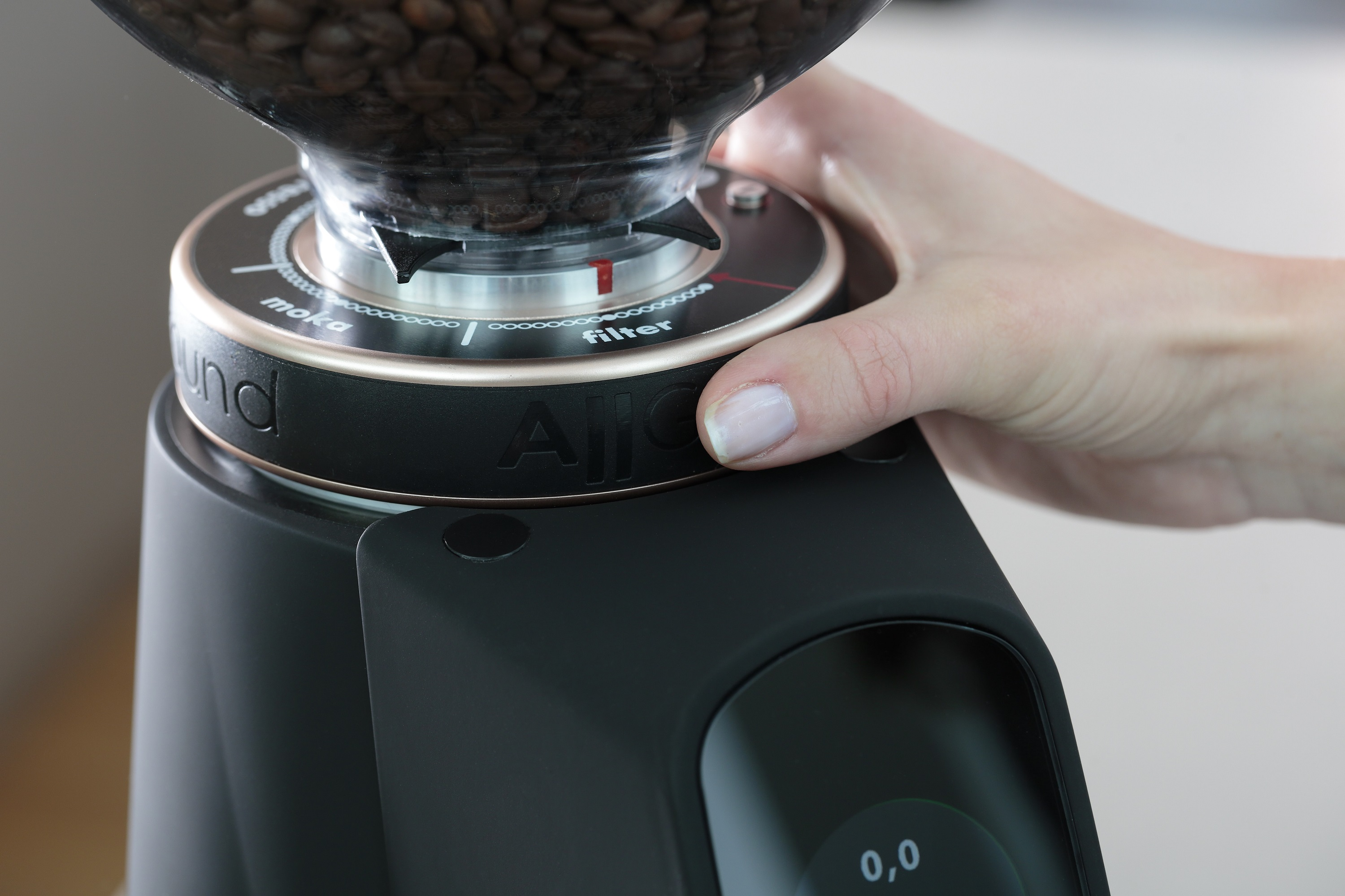 Eine Hand, die die Mahleinstellung an einer schwarzen Kaffeemühle einstellt. Kaffeebohnen oben.