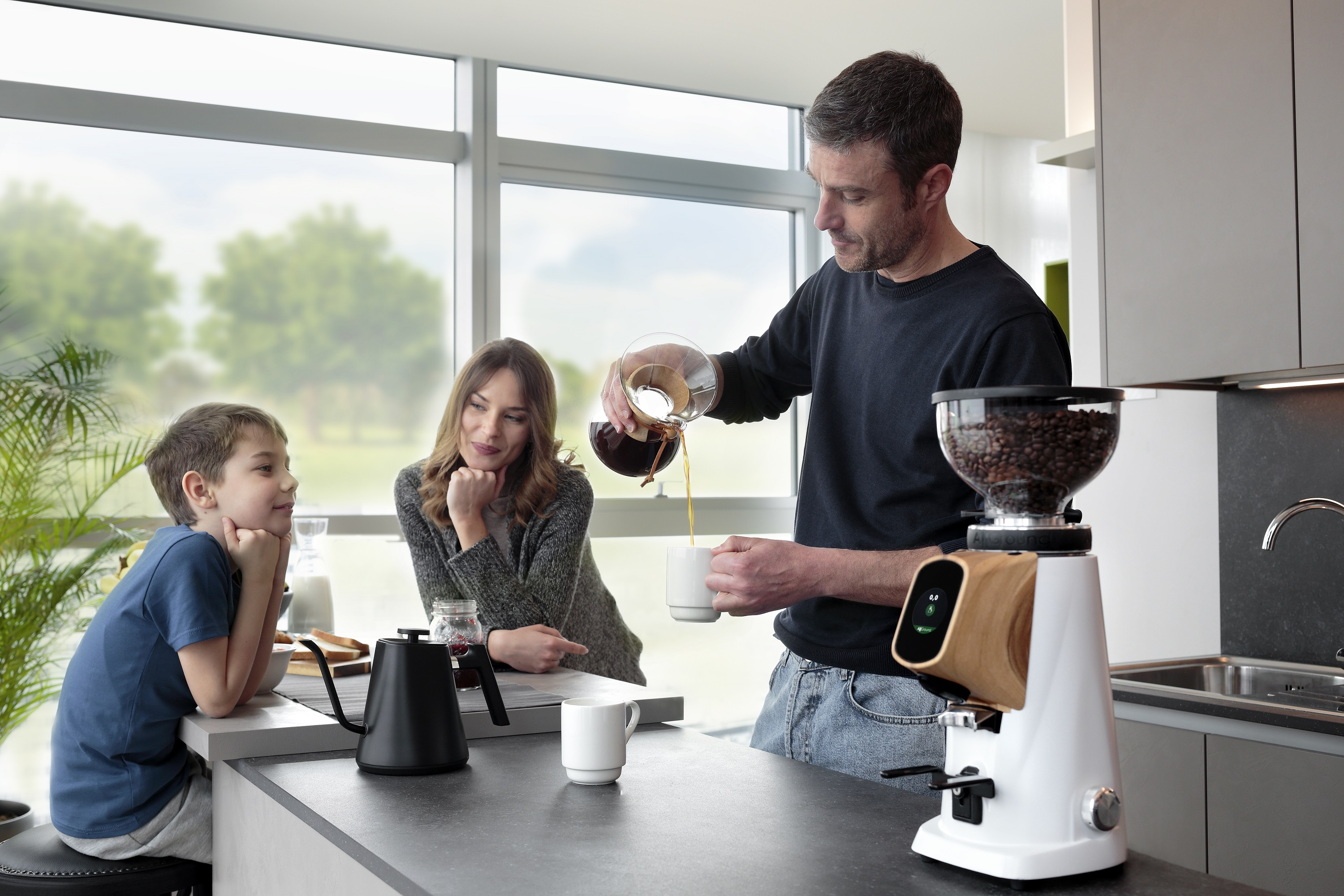 Ein Mann gießt Kaffee in eine Tasse, während eine Frau und ein Junge in einer Küche zusehen.