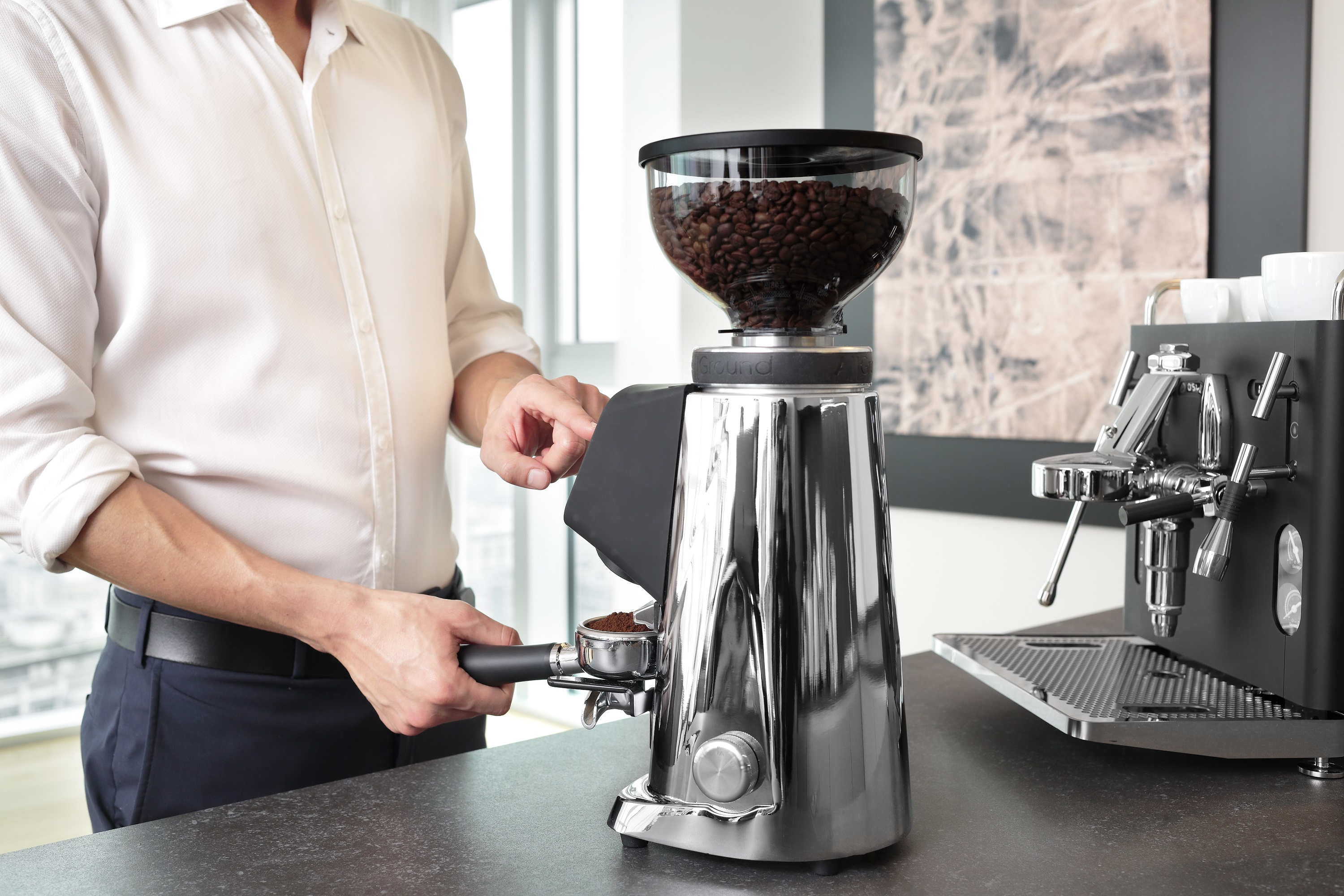 Ein Mann mahlt Kaffeebohnen mit einer Chrom-Kaffeemühle, in der Nähe einer Espressomaschine.
