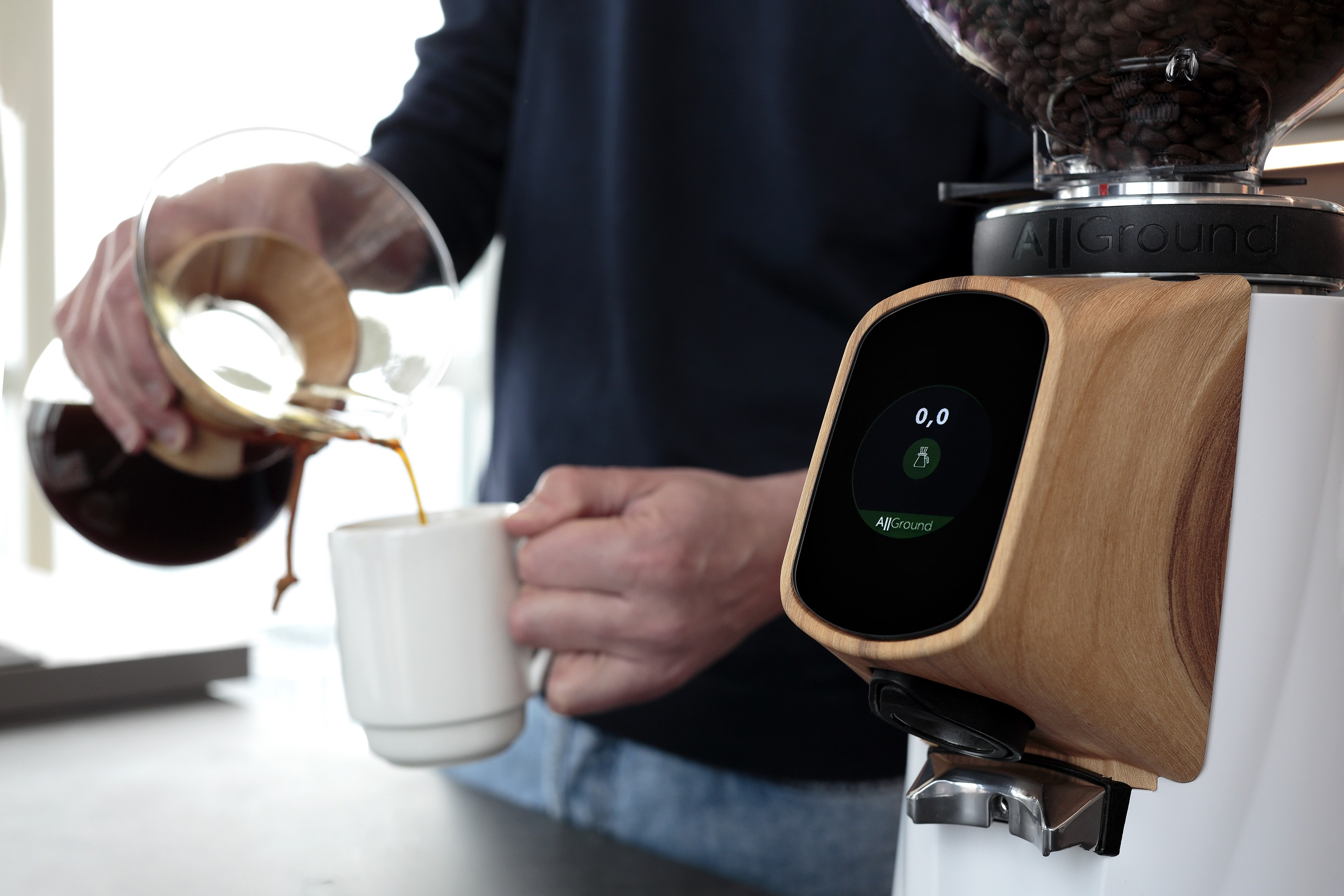 Eine Person gießt Kaffee aus einem Glasbehälter in eine weiße Tasse. Kaffeemaschine rechts.