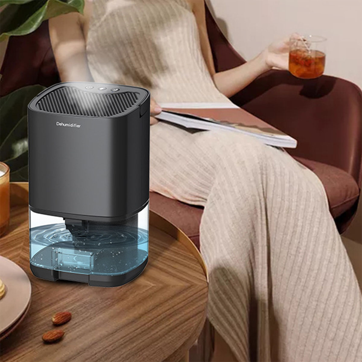 Osuszacz z zbiornikiem na wodę stoi na stole. Obok siedzi osoba trzymająca szklankę.