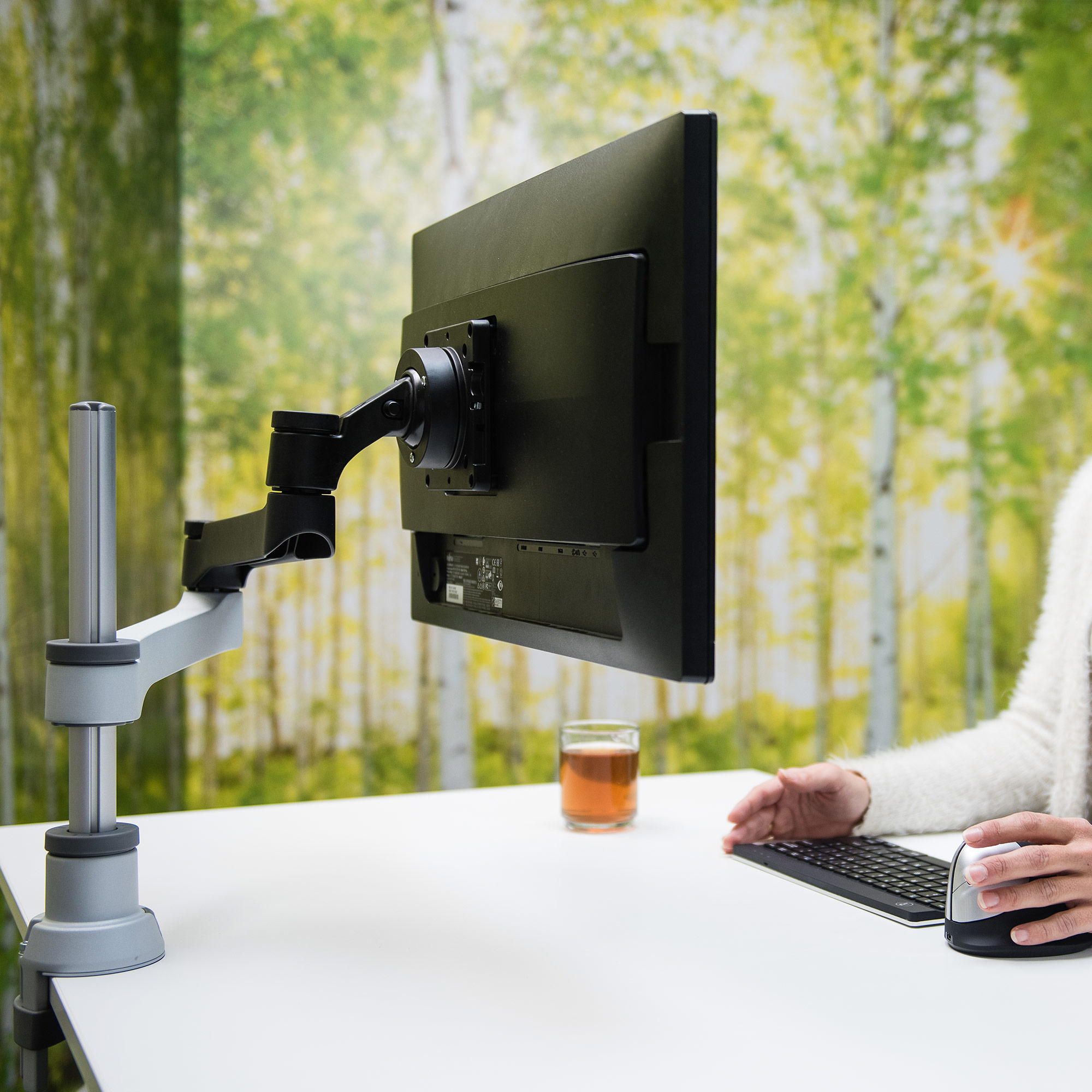 Un moniteur d'ordinateur monté sur un bureau. Une personne utilise un clavier et une souris avec un fond de forêt.