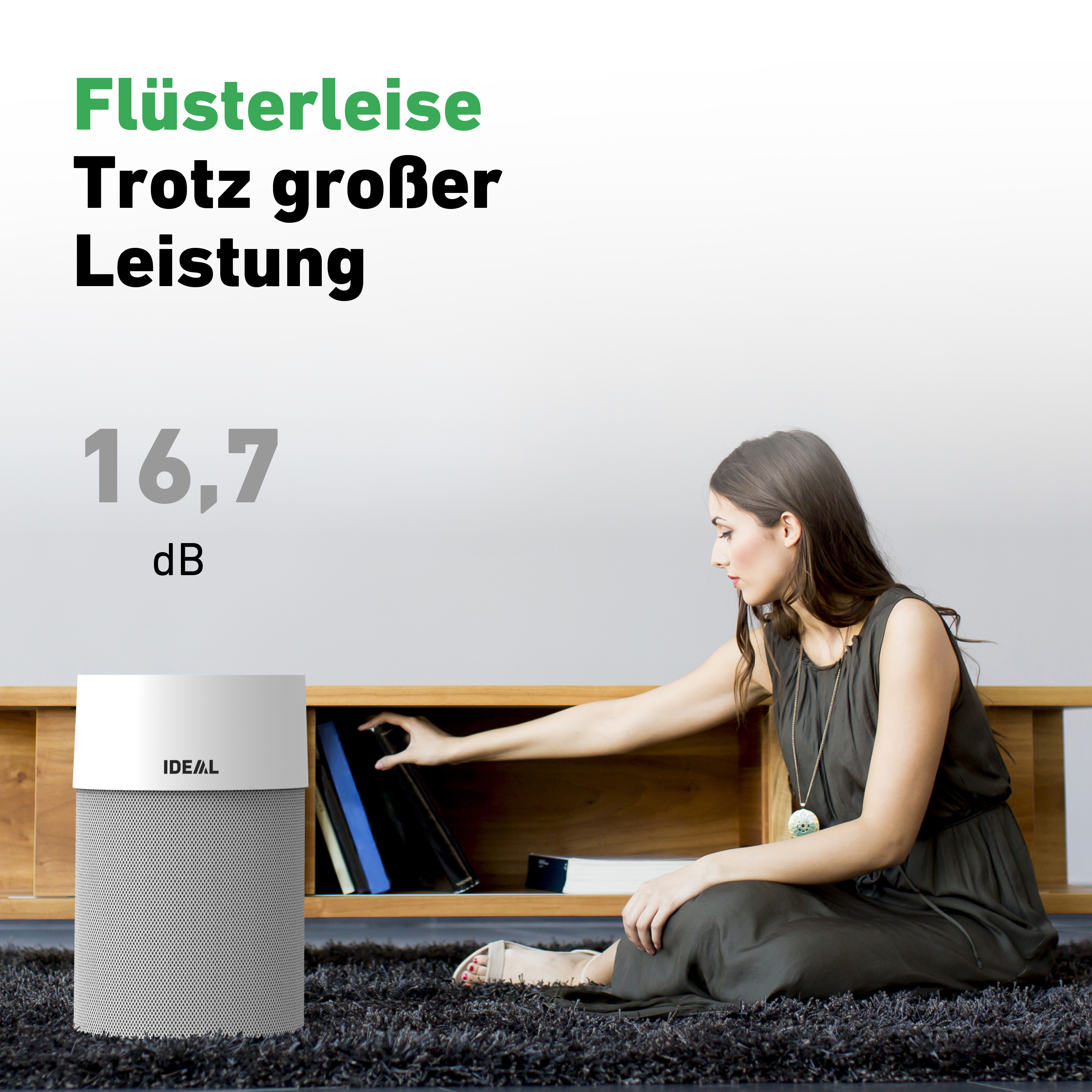 Eine Frau sitzt neben einem weiß-grauen Luftreiniger und greift nach einem Stapel Bücher auf einem Holzregal.