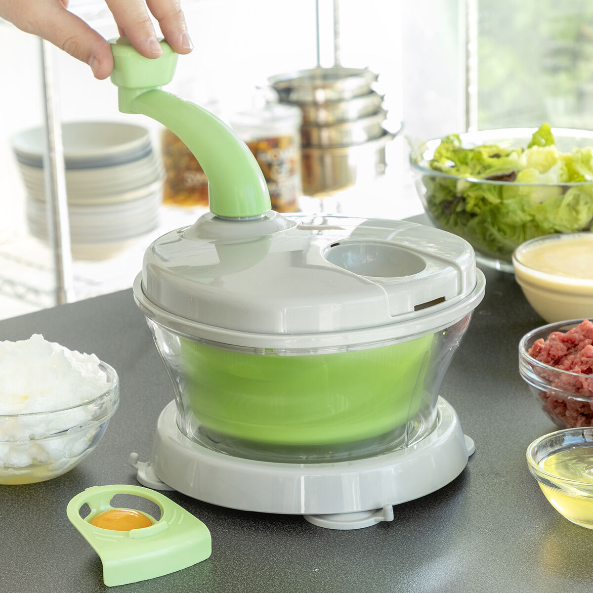 Un tritatutto da cucina verde e bianco con ingredienti intorno e una mano che gira la maniglia.