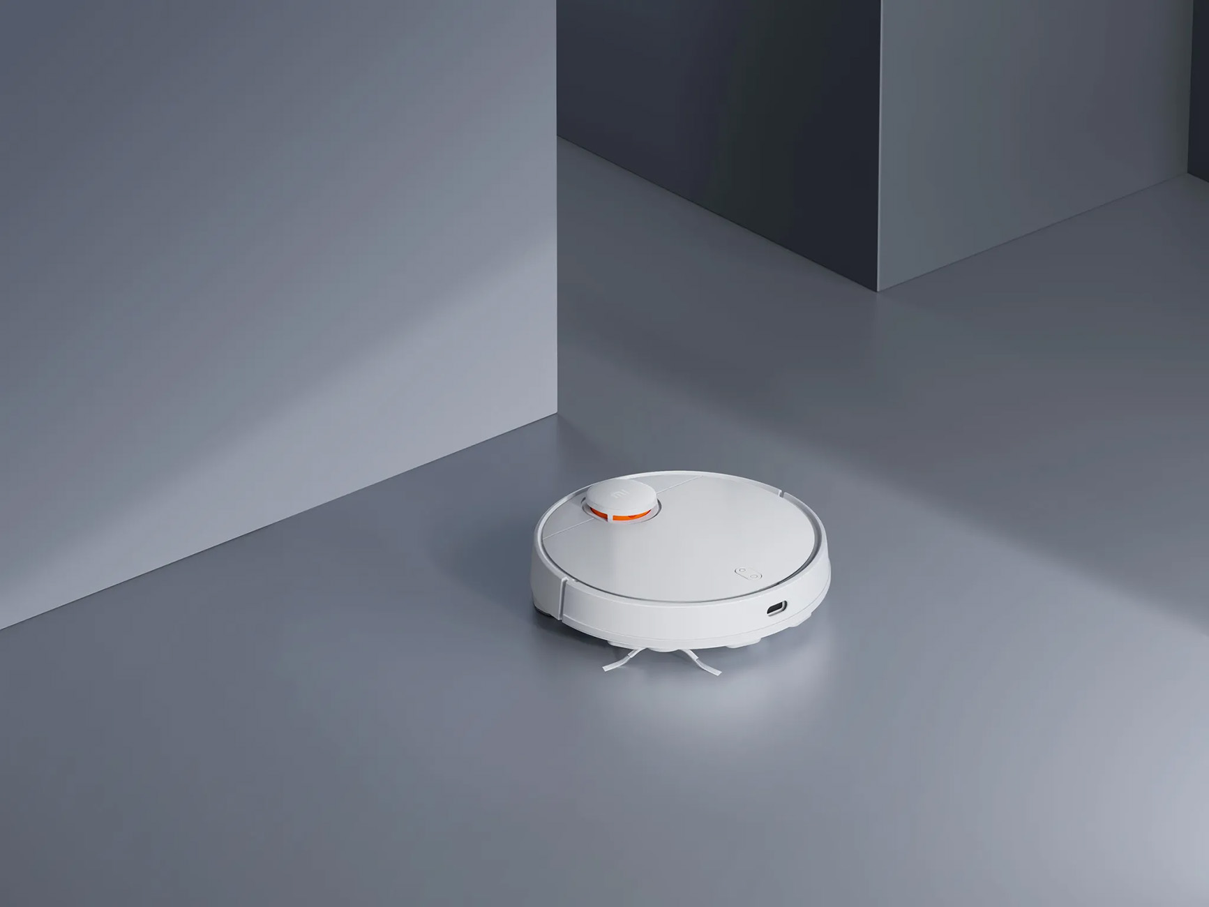 Ein weißer Roboter-Staubsauger auf grauem Boden. Er hat einen orangefarbenen Knopf und einige Bürsten.