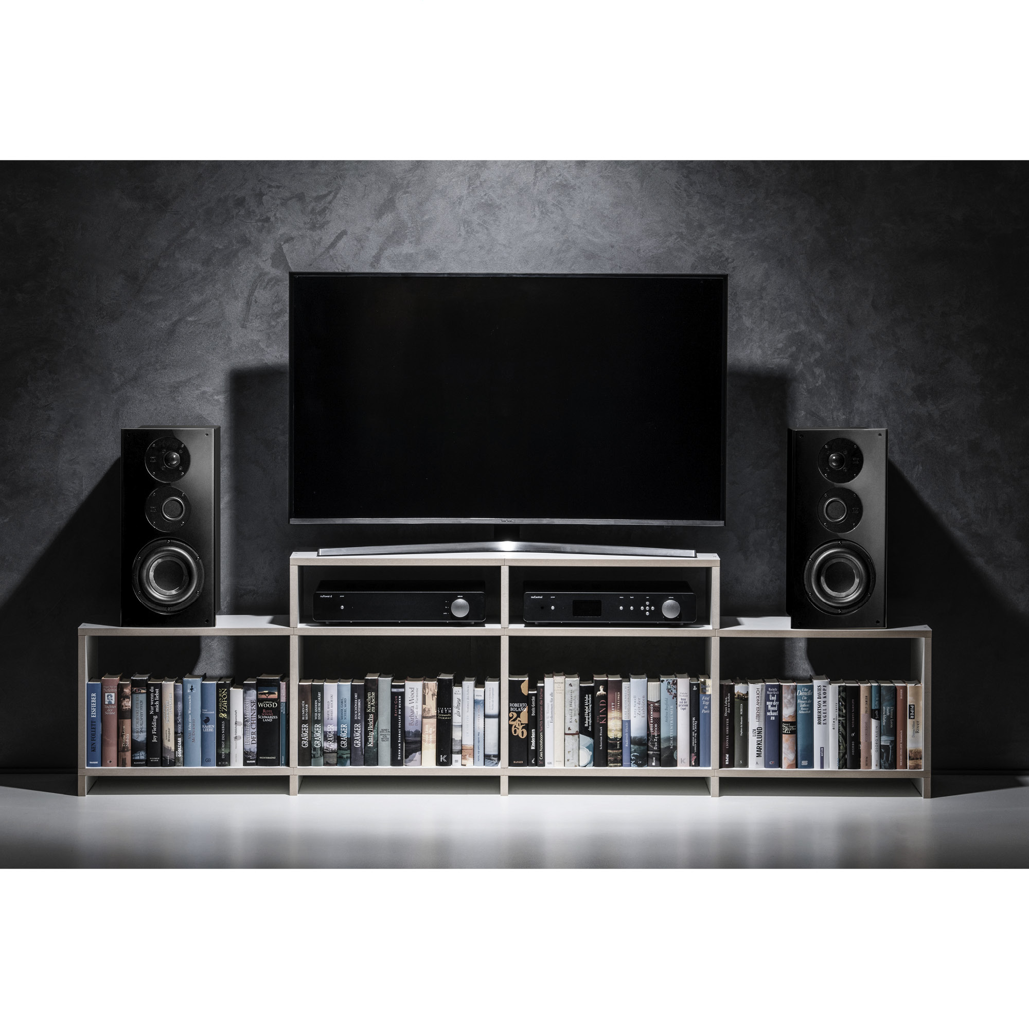 Moderne Wohnzimmereinrichtung mit Fernseher auf weißem Regal, flankiert von schwarzen Lautsprechern, Büchern und dunkler Wand.