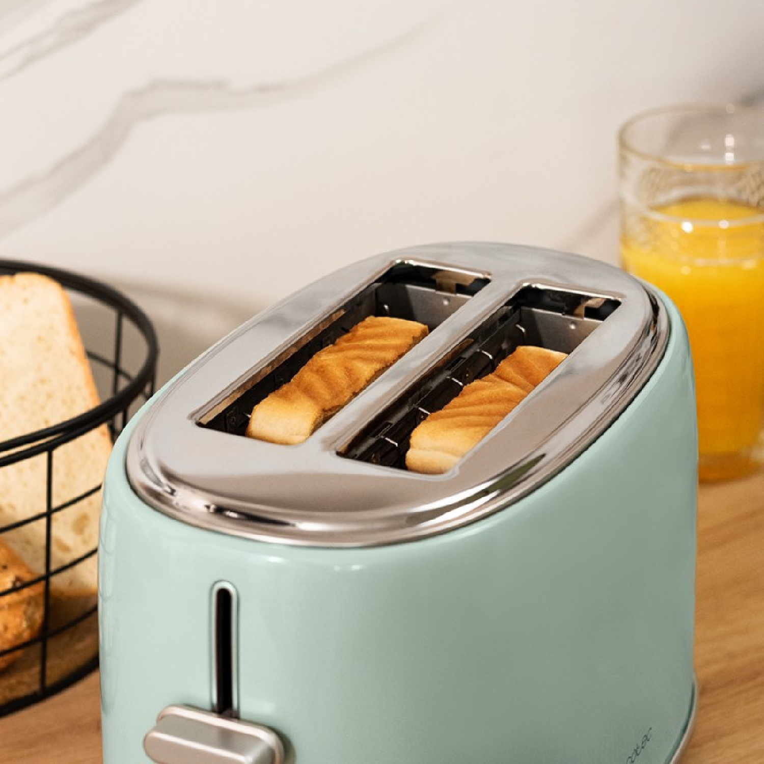 Mint-green toaster with two slots. Toast is in the slots, and juice and bread are nearby.