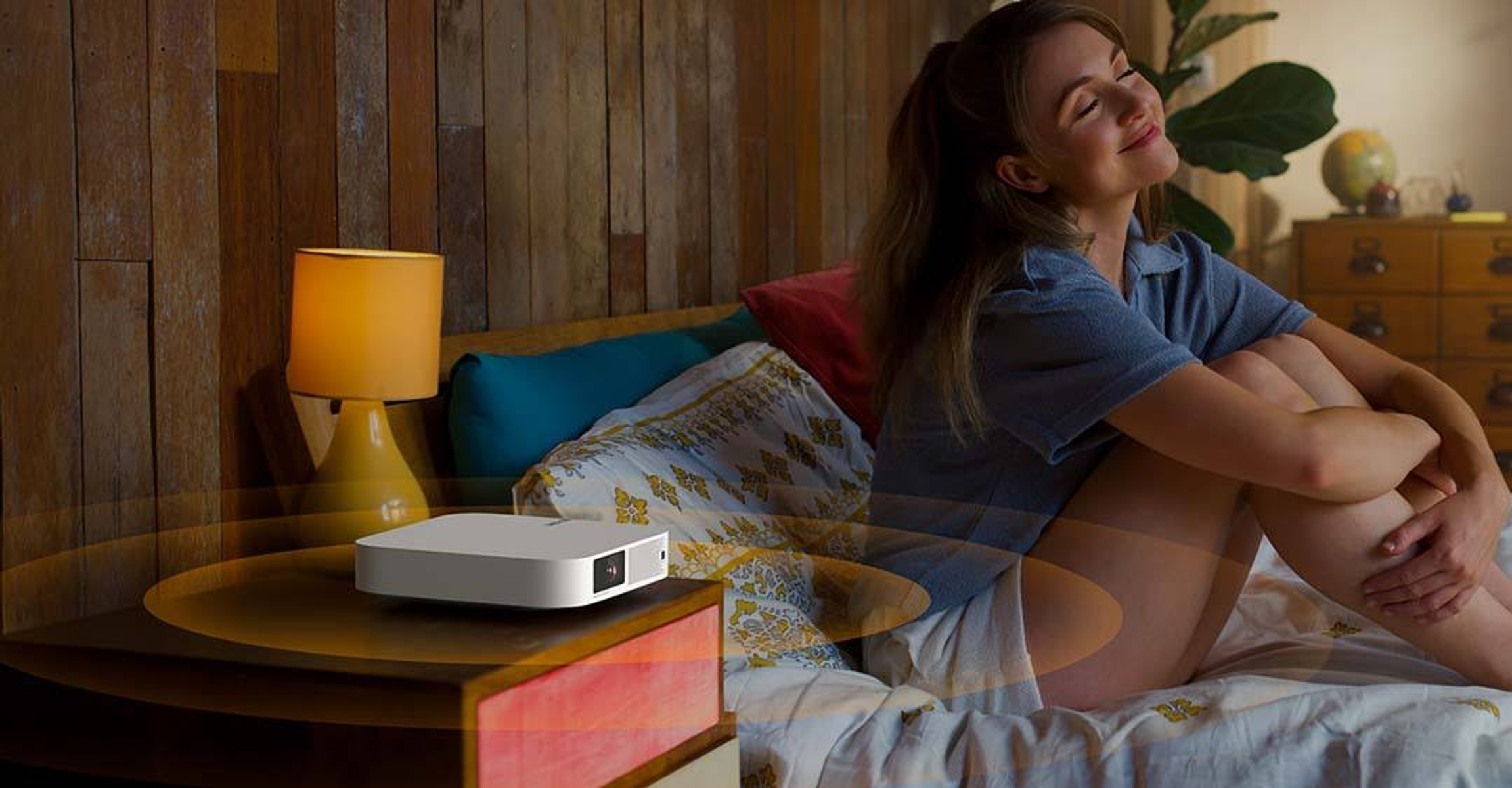 Frau im Bett mit kleinem Projektor, warmem Licht und weißem Gerät.