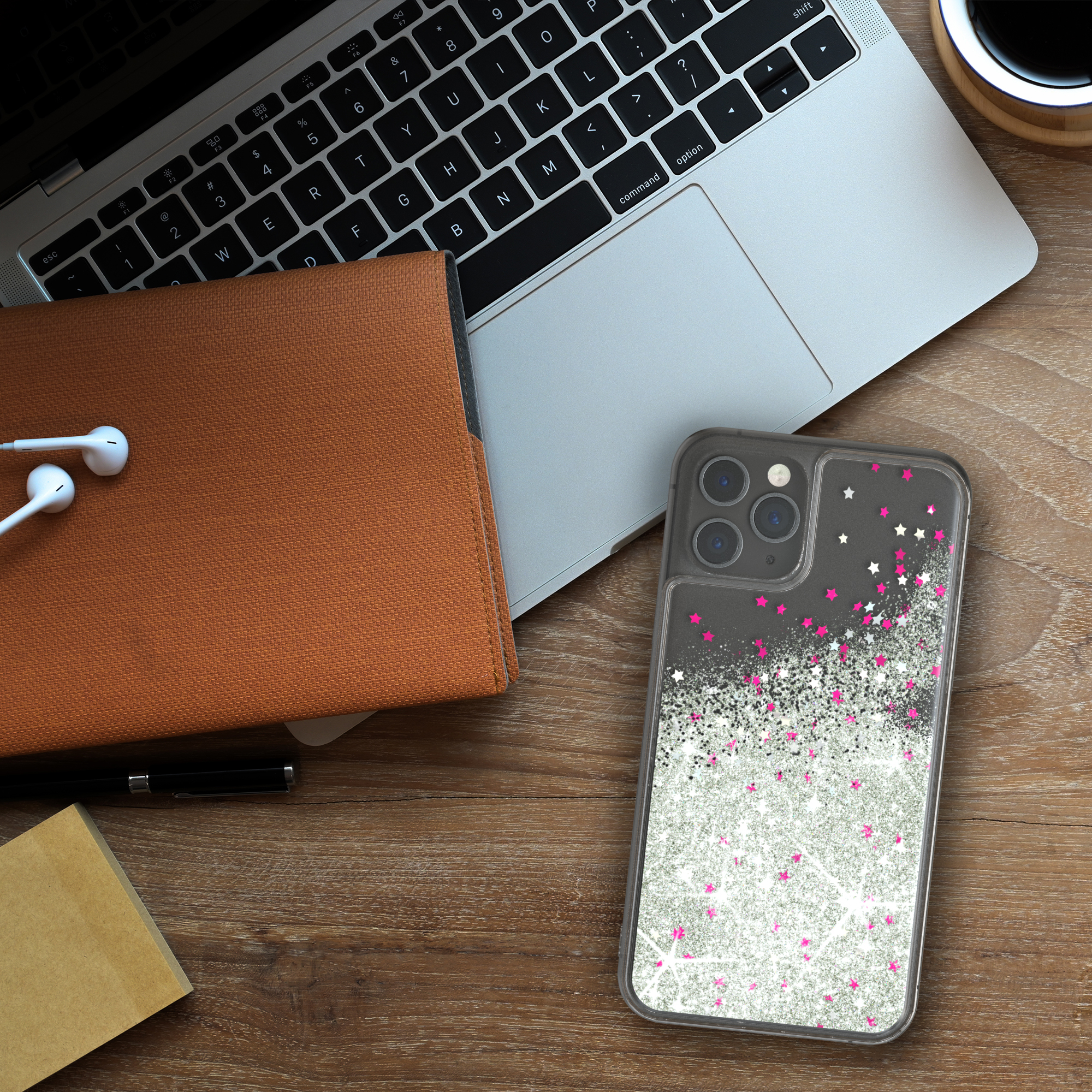 Laptop, Notizbuch, Stift und Telefon mit Glitzerhülle auf dem Holztisch. Weiße Kopfhörer daneben.