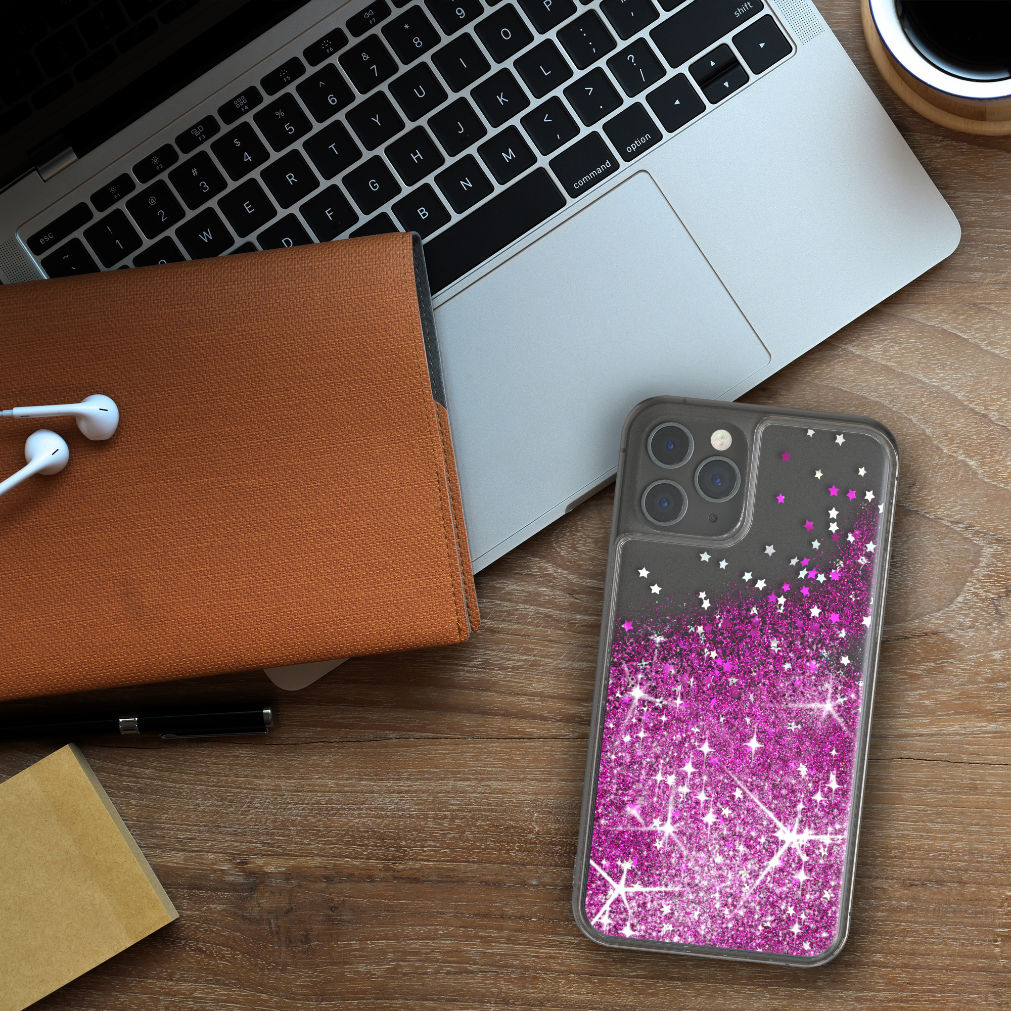 Laptop, Telefon mit pinkfarbener Glitzerhülle, Notizbuch und Kopfhörer auf einem Holztisch.