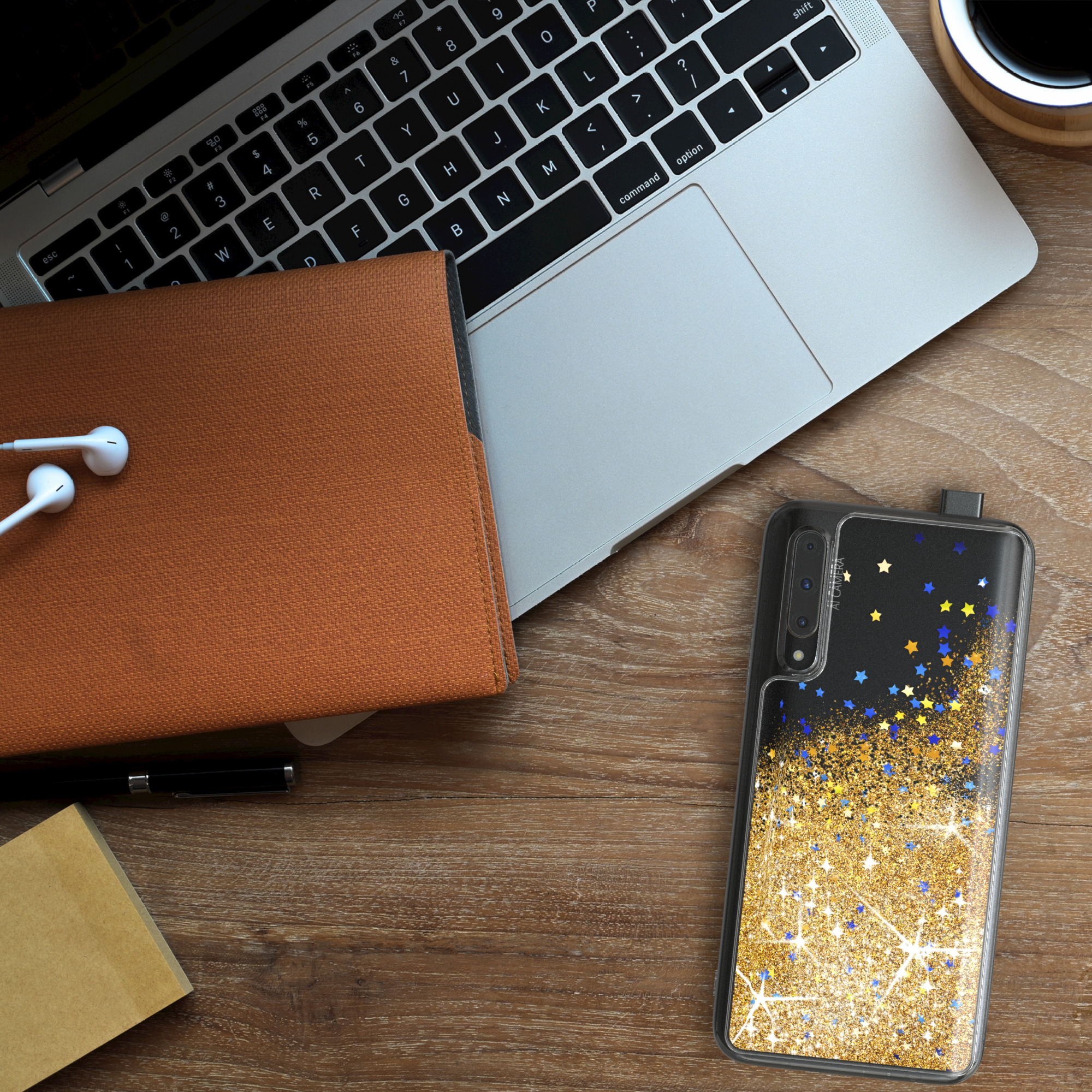 Laptop, Handyhülle mit Goldglitter, Notizbuch und Kopfhörer auf einem Holztisch.