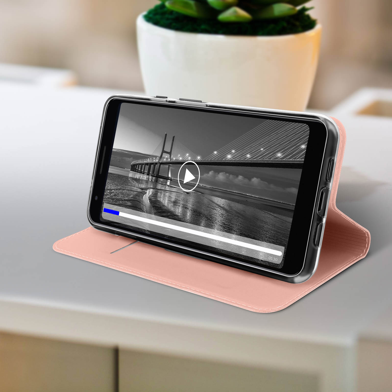 Ein Smartphone zeigt ein Brückenvideo, das auf einer rosa Hülle auf einer grauen Oberfläche liegt, mit einer Pflanze im Hintergrund.