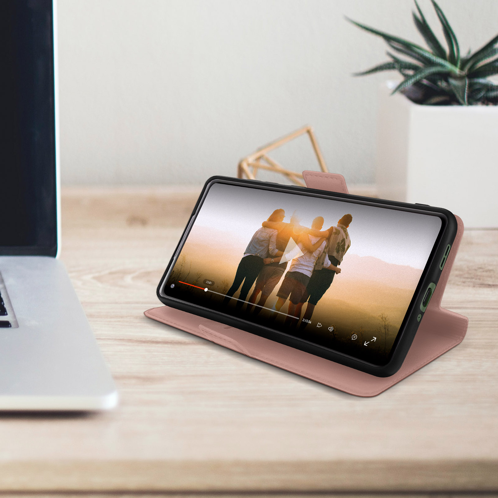 Telefono in custodia rosa su un tavolo di legno, che riproduce un video. Un laptop e una pianta sullo sfondo.