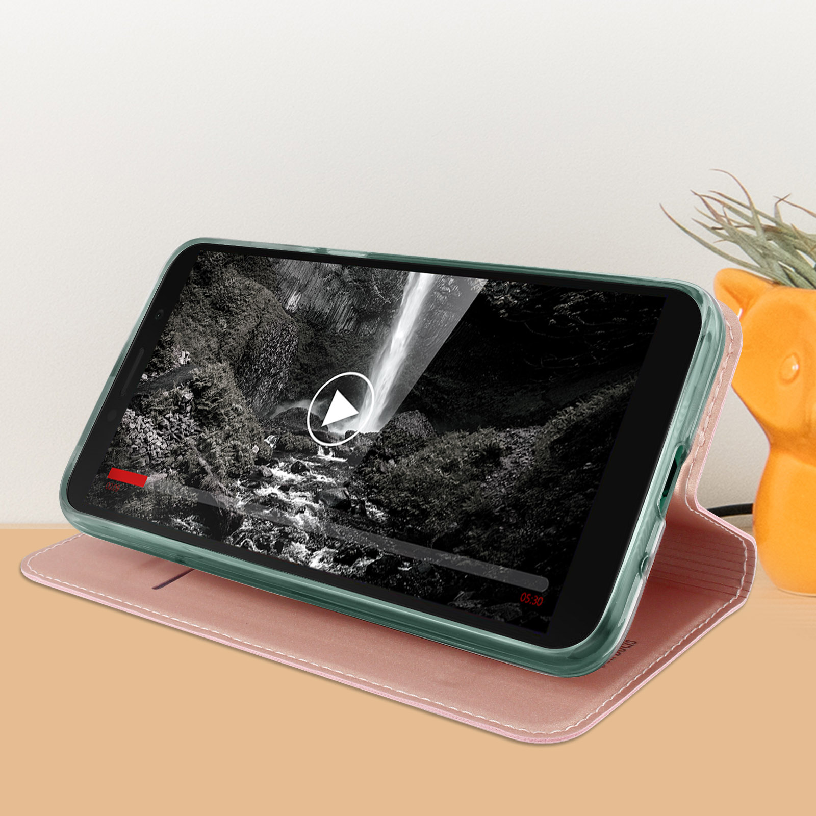 Smartphone in custodia rosa, riproduce video di una cascata. Vaso di fiori sullo sfondo.
