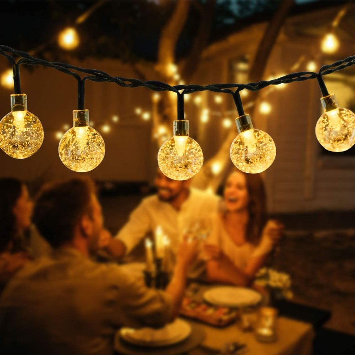 Eine Lichterkette im Freien mit verschwommenen Personen im Hintergrund, bei einer Dinnerparty.