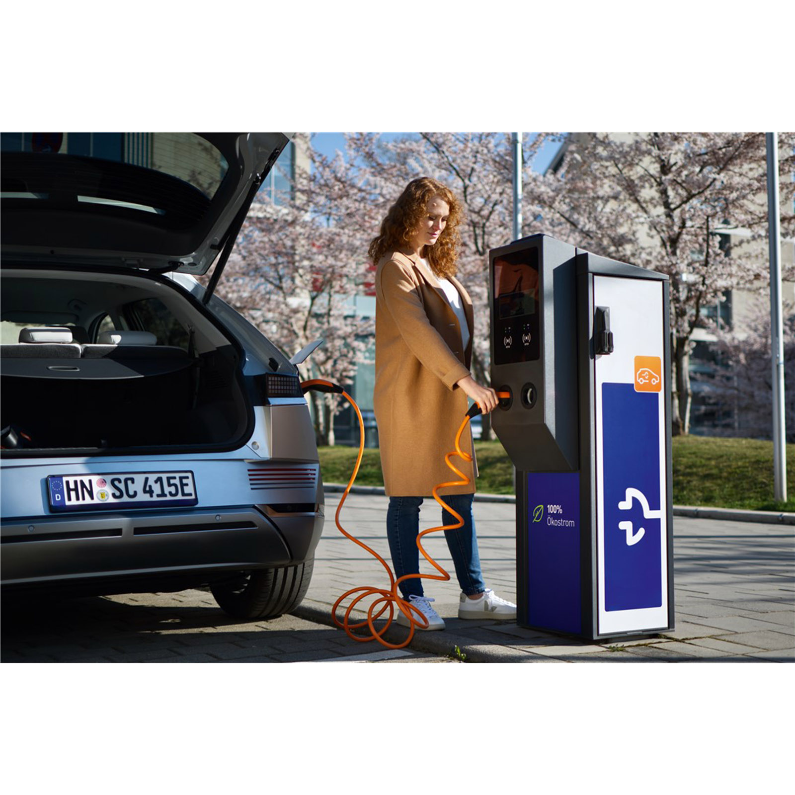 Eine Frau, die ein Elektroauto auflädt. Das Auto ist silberfarben und die Ladestation ist blau und weiß.