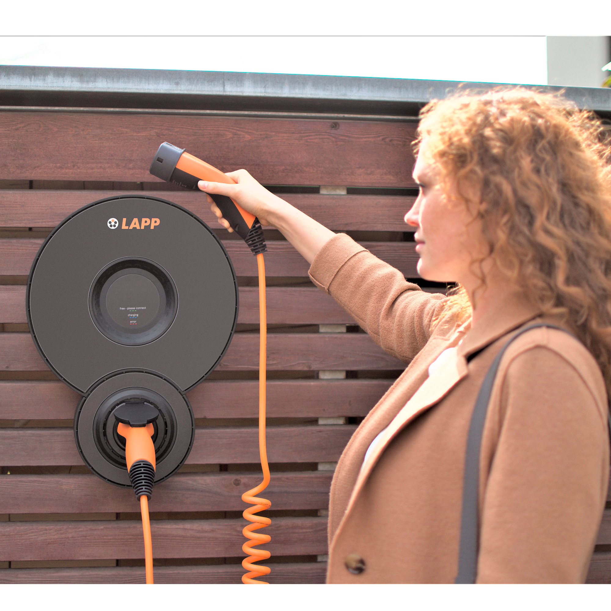 Eine Frau lädt ein Elektroauto mit einer Ladestation an einem Holzzaun auf.