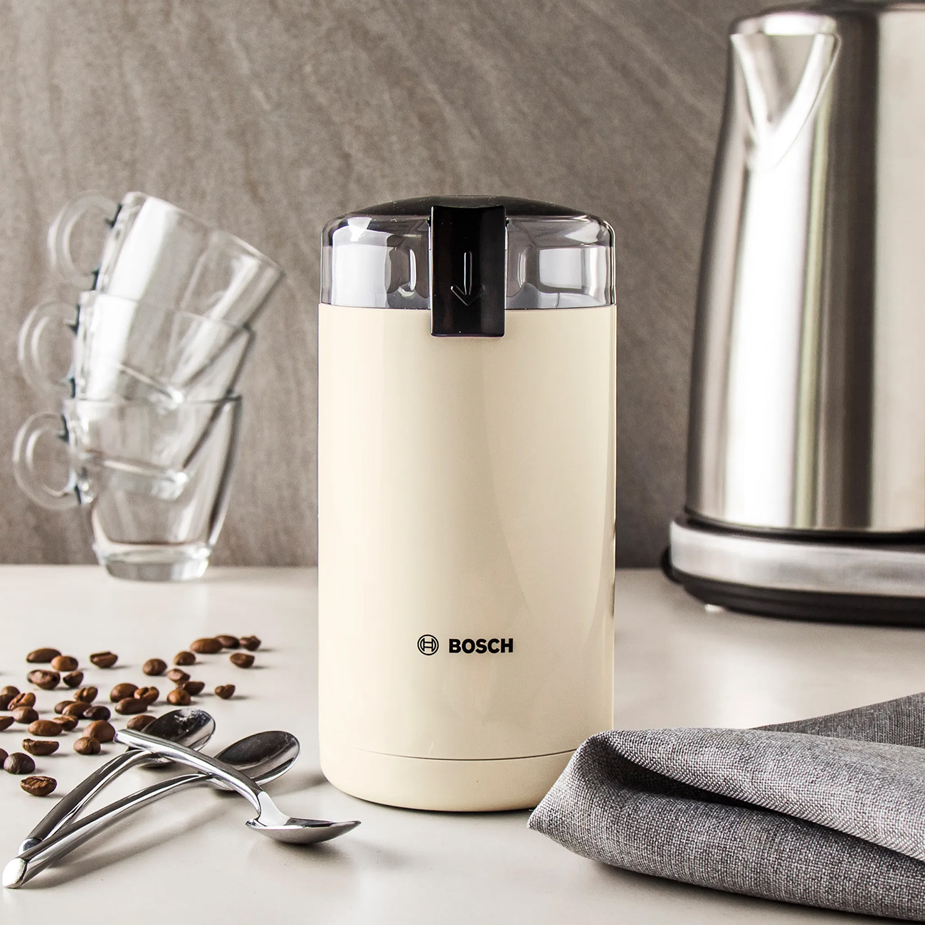 Kremowy młynek do kawy z czarną pokrywką, obok ziaren kawy, łyżek i czajnika.