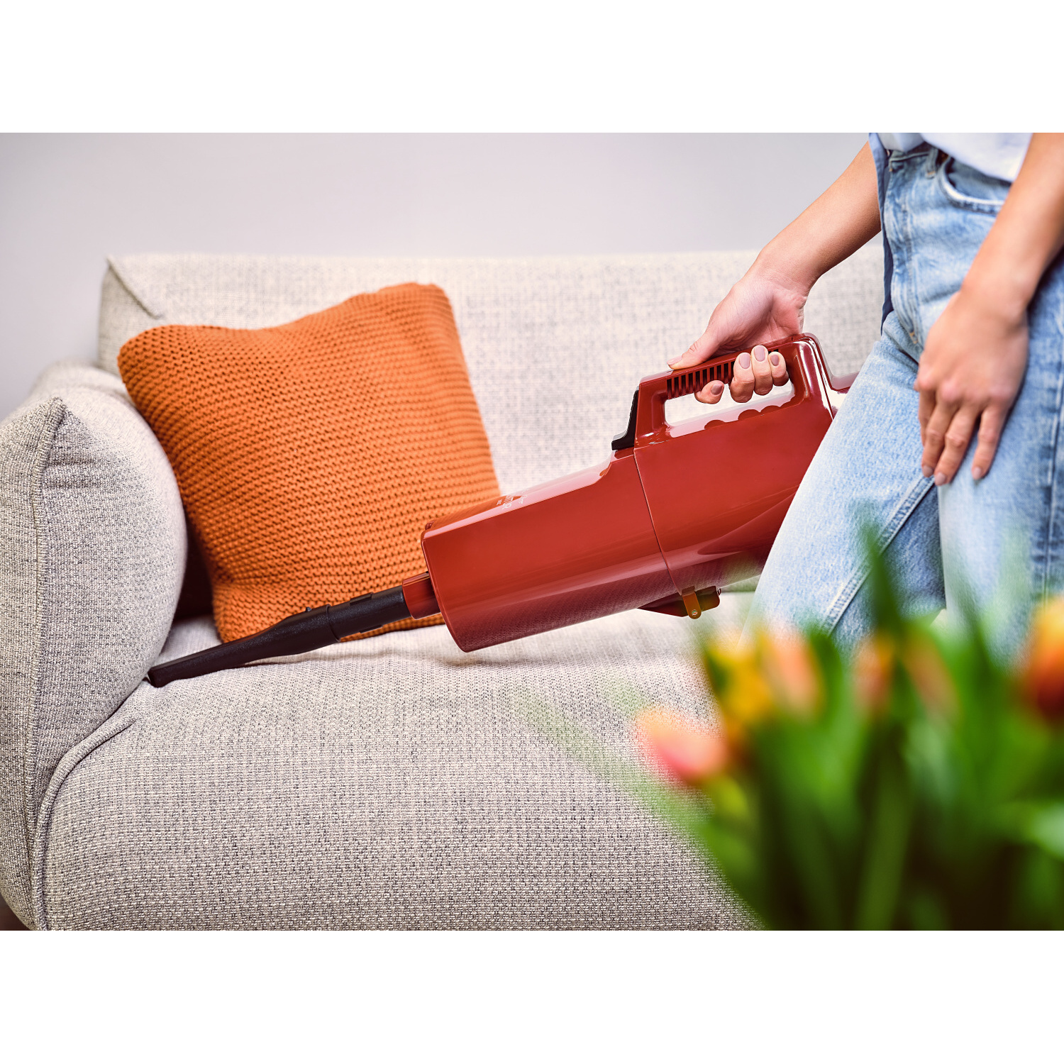Person verwendet roten Handstaubsauger auf grauer Couch, mit orangefarbenem Kissen und Blumen.