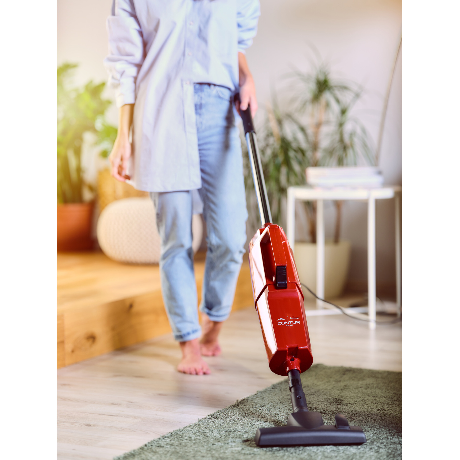 Person saugt einen Raum mit einem roten Staubsauger. Der Boden ist grüner Teppich und Holz.