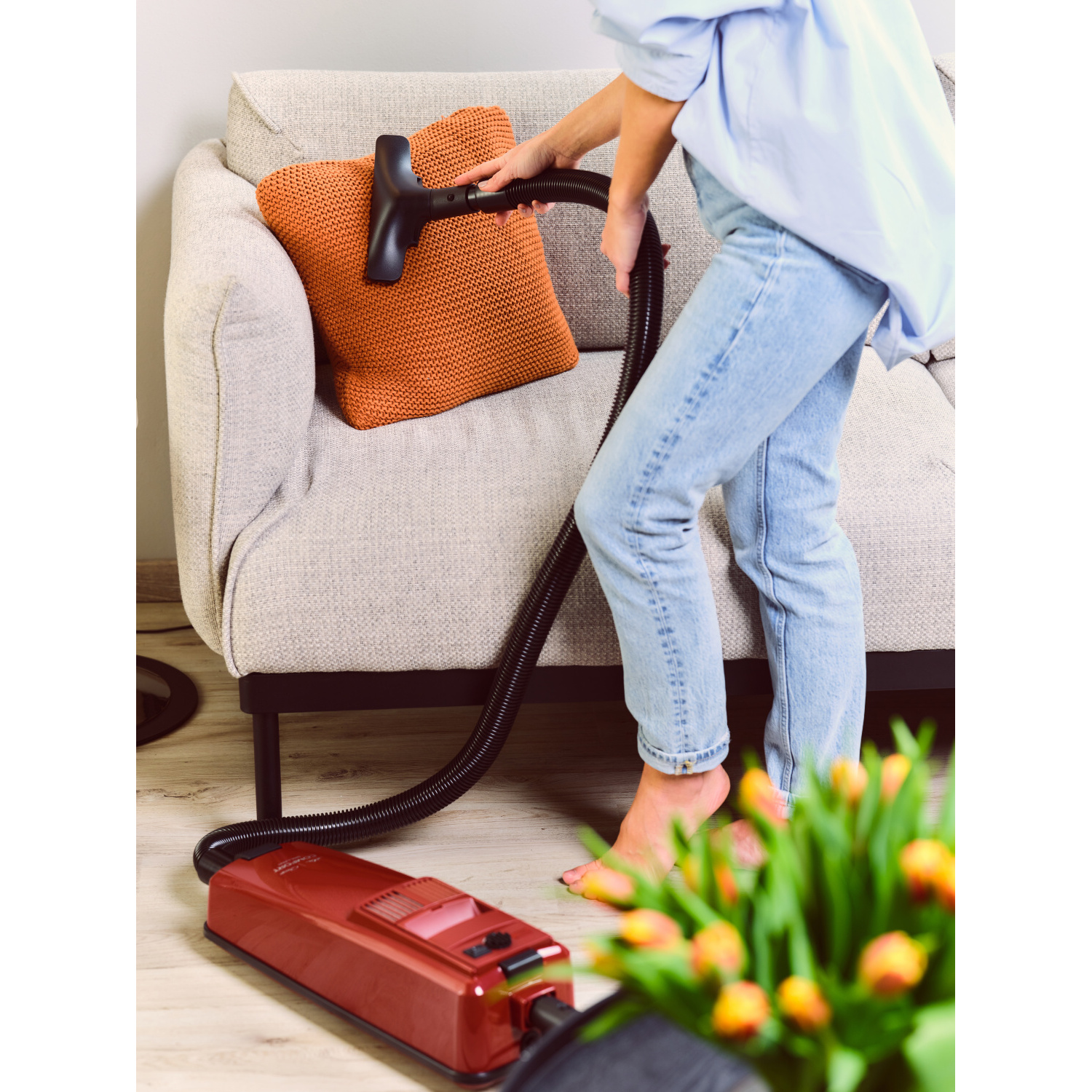 Eine Person saugt ein Sofa mit einem orangefarbenen Kissen. Ein Strauß gelber Blumen im Vordergrund.