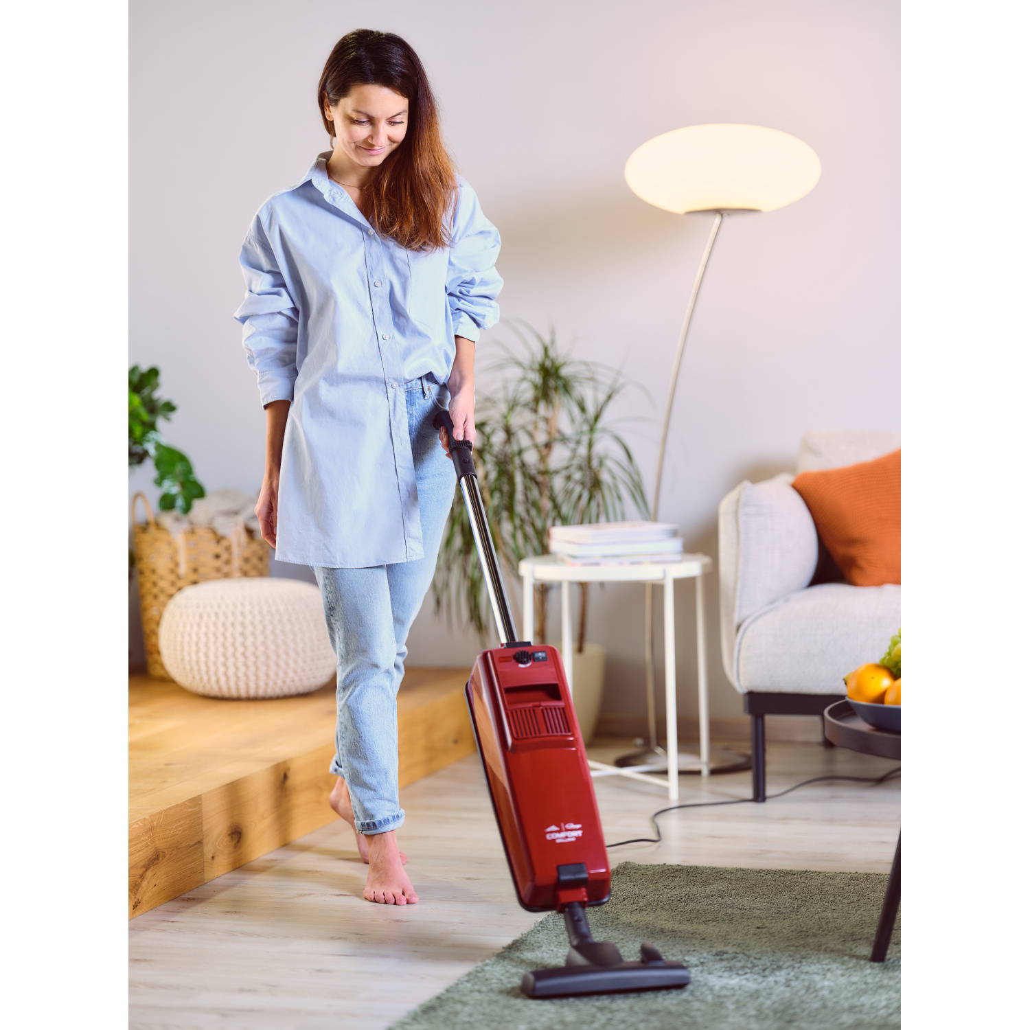 Eine Frau saugt einen Raum mit einem roten Staubsauger. Sie trägt ein blaues Hemd und Jeans.