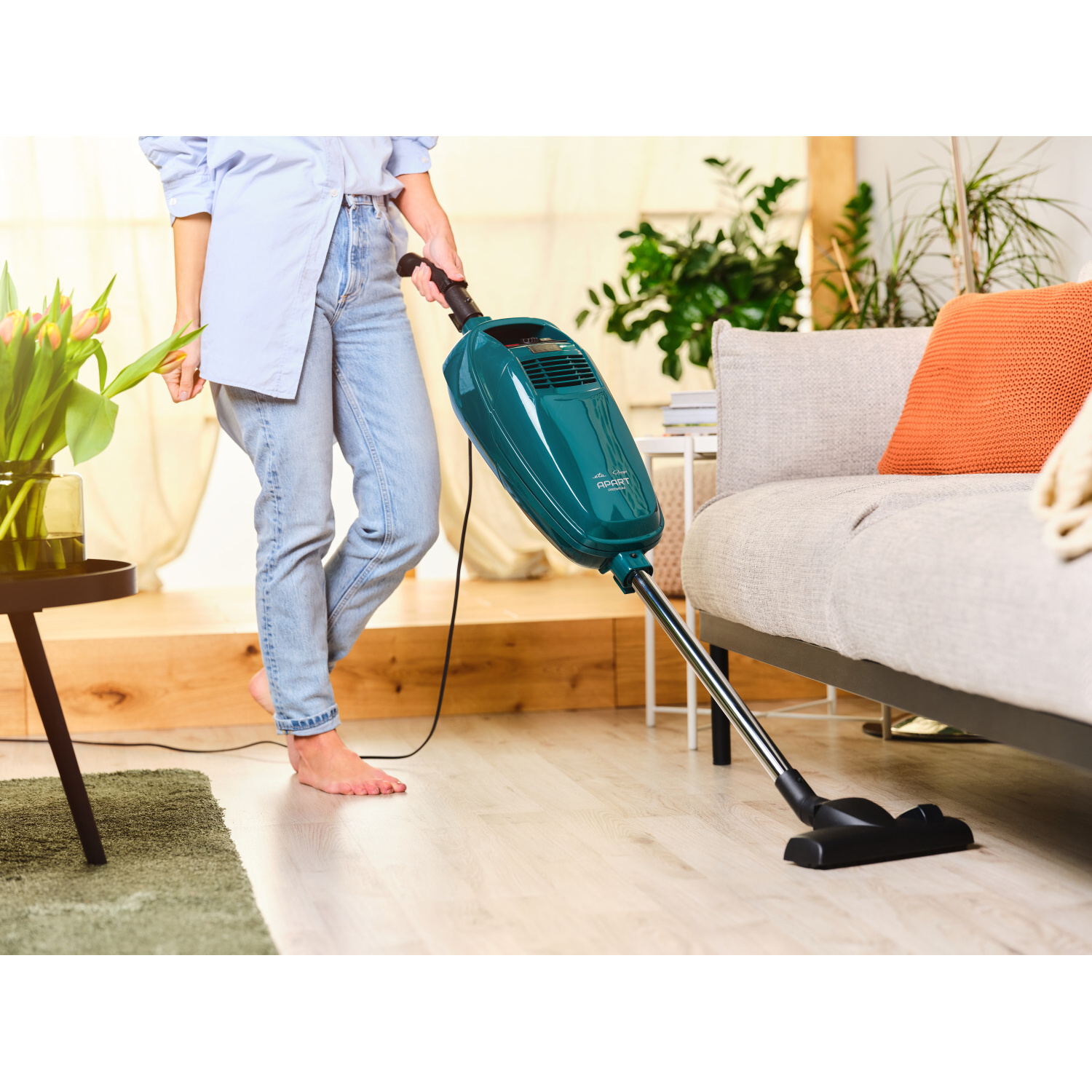Person saugt den Boden mit einem grünen Staubsauger in der Nähe von Couch und Blumen.