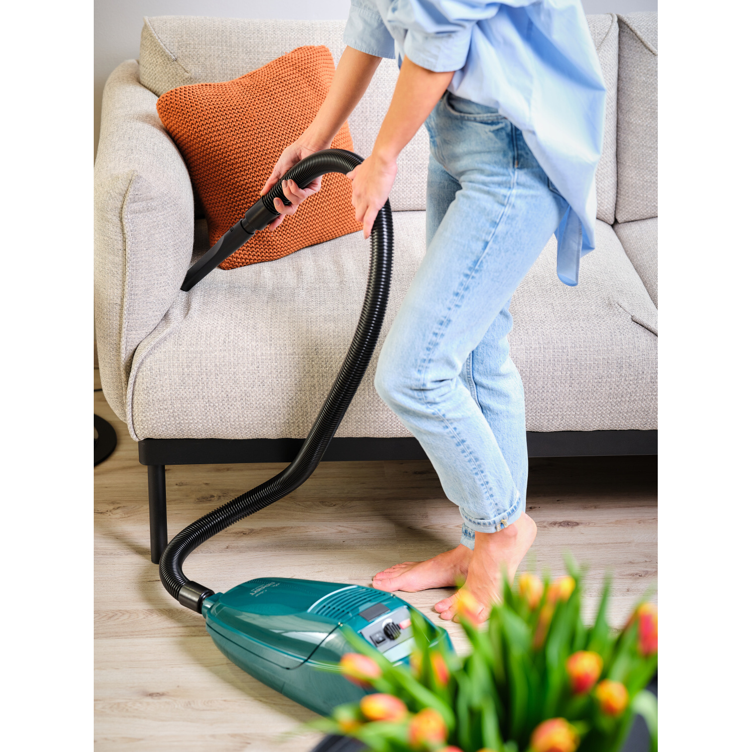 Eine Person saugt die Couch mit einem grünen Staubsauger und einem schwarzen Schlauch ab.