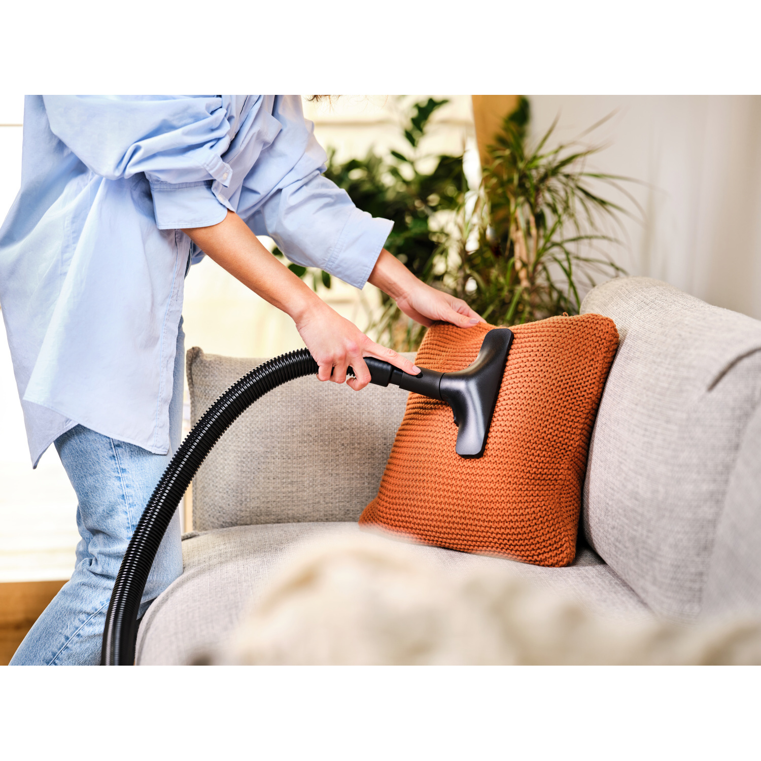 Eine Person, die einen orangenen Kissen auf einem grauen Sofa mit einem Staubsauger absaugt.